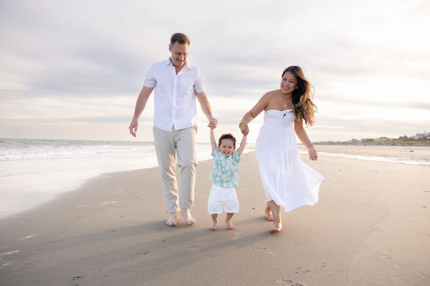 Happy family walking on beach, parents holding toddler's hands, wearing white outfits, ocean waves in background.