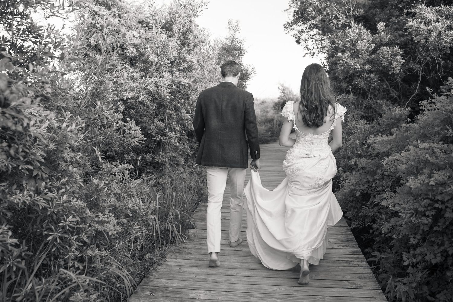 Black and white photo of a bride and groom walking hand in hand on a wooden boardwalk surrounded by lush greenery.