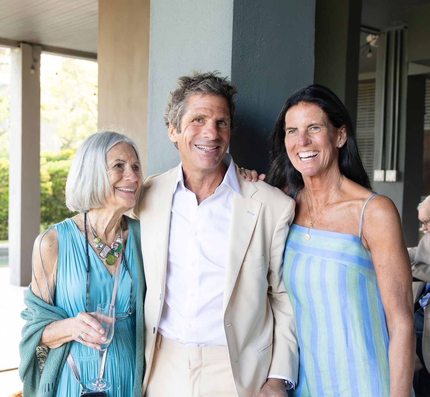 Three adults pose together smiling at outdoor wedding reception on a sunny day.