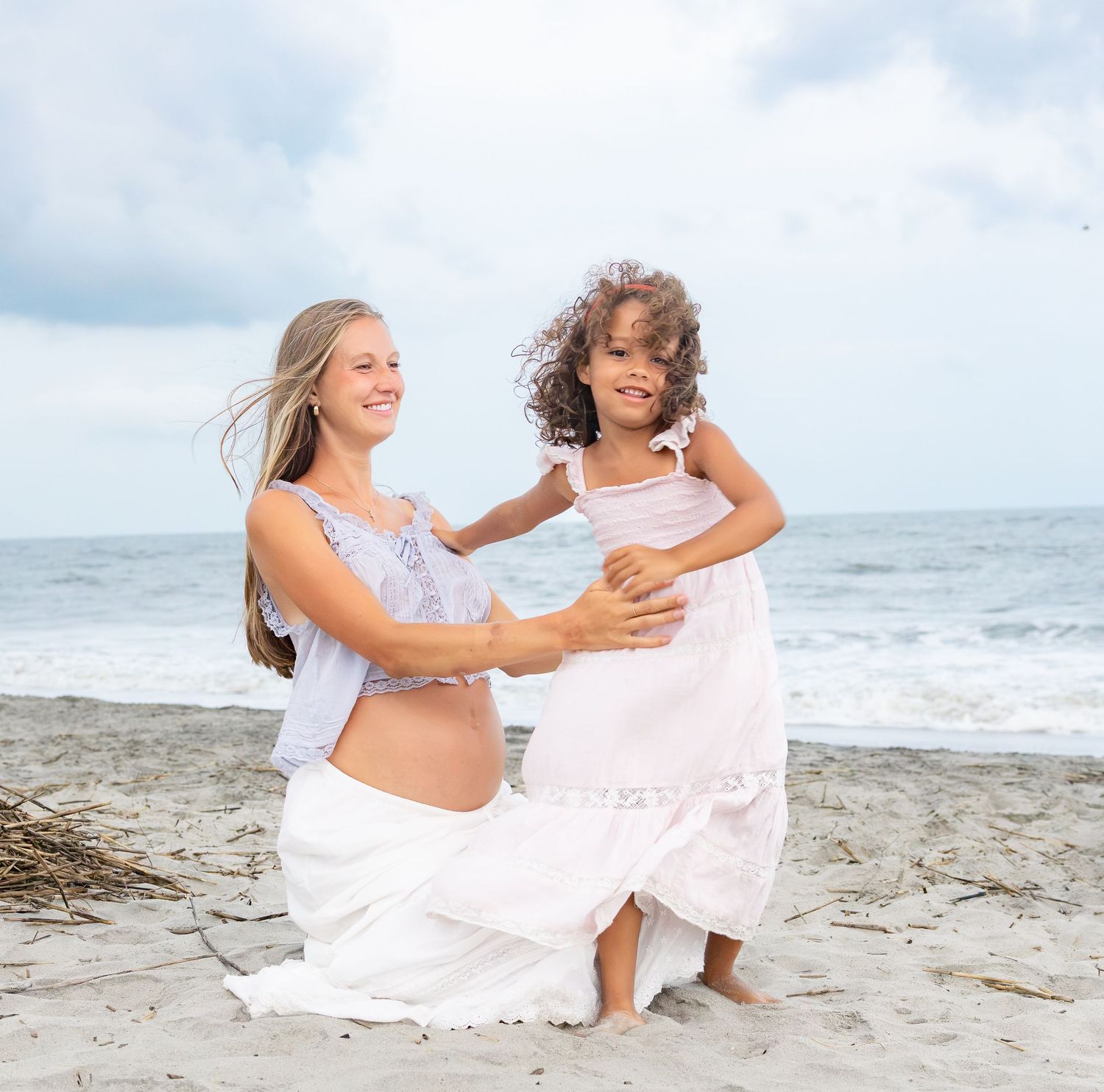 Pregnant woman and young girl laughing together on a sandy beach, wearing white and pink dresses by the ocean.