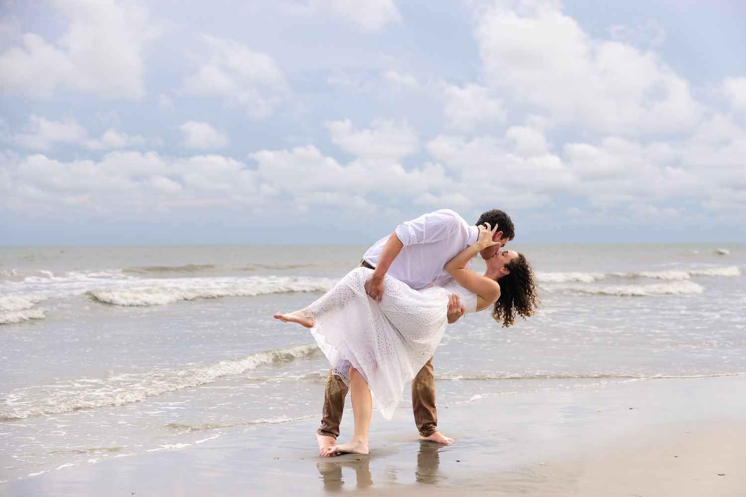 Romantic couple sharing a passionate kiss on a sandy beach with ocean waves and cloudy blue sky backdrop.