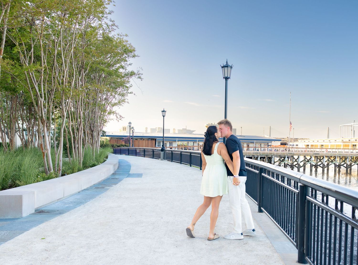 Couple sharing a romantic kiss on a waterfront promenade with a pier and lamp post in the background.