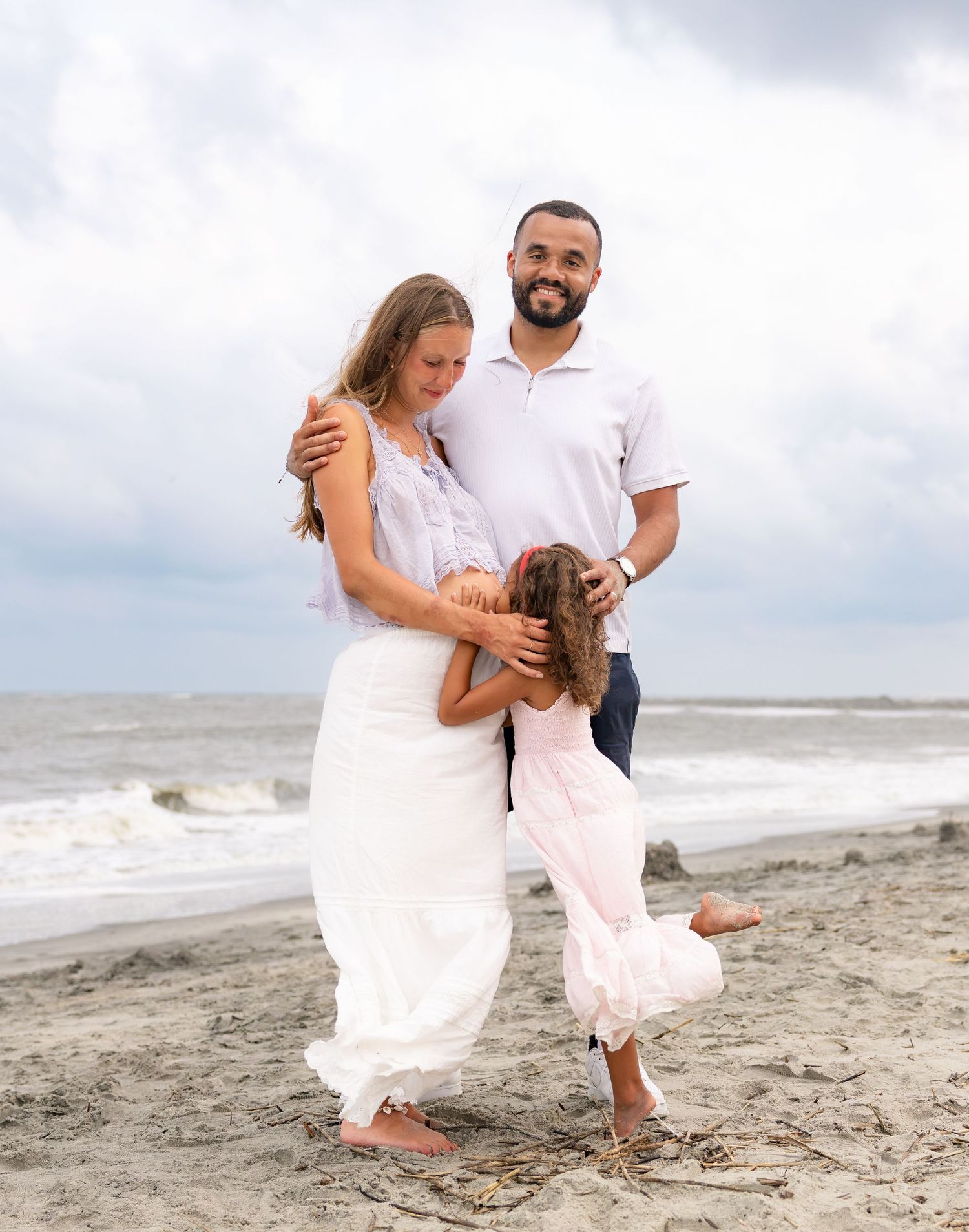 Family of three posing on a sandy beach, parents embracing while young daughter hugs mom, ocean waves in background.