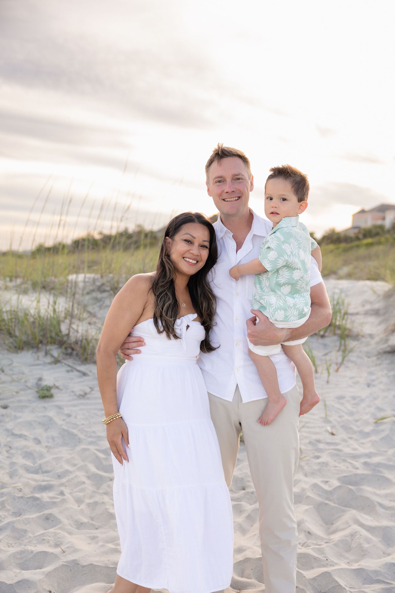 Happy family of three posing on a sandy beach at sunset, parents in white outfits holding their baby boy.
