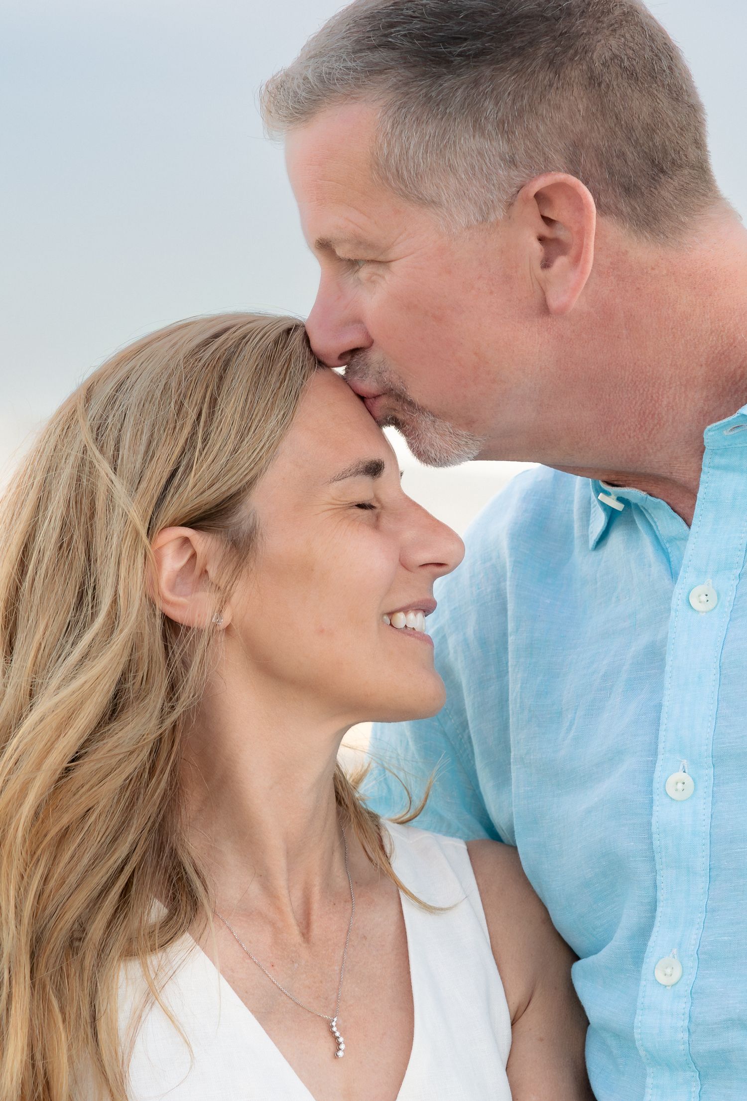 A middle-aged couple shares a tender moment at the beach, man kissing woman's forehead as she smiles warmly.