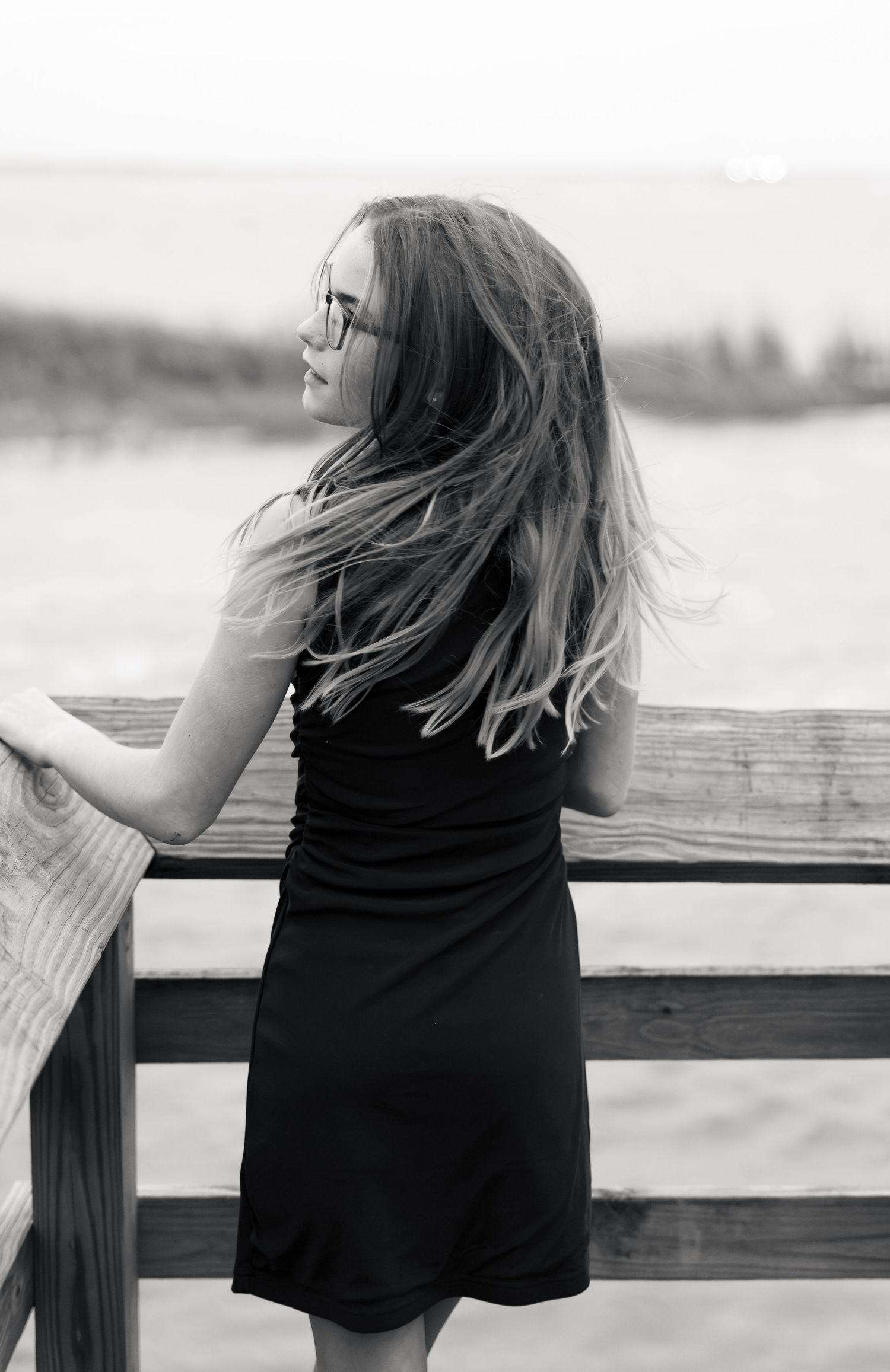 Black and white photo of a woman in glasses and a black dress, hair blowing in the wind, leaning on a wooden pier railing above the water.