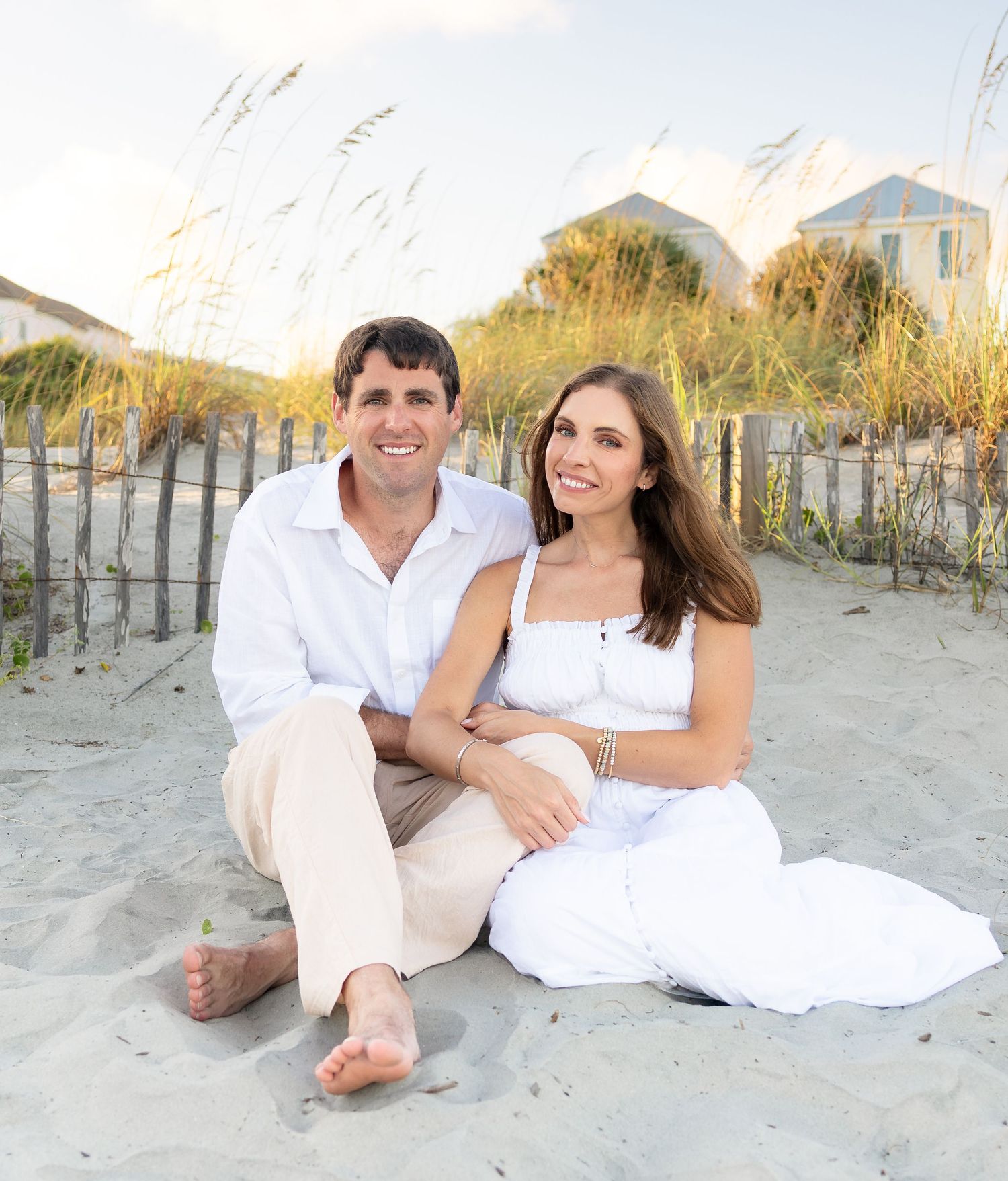 Smiling couple in white outfits sitting barefoot on a sandy beach dune with sea oats, a wooden fence, and coastal homes behind them.