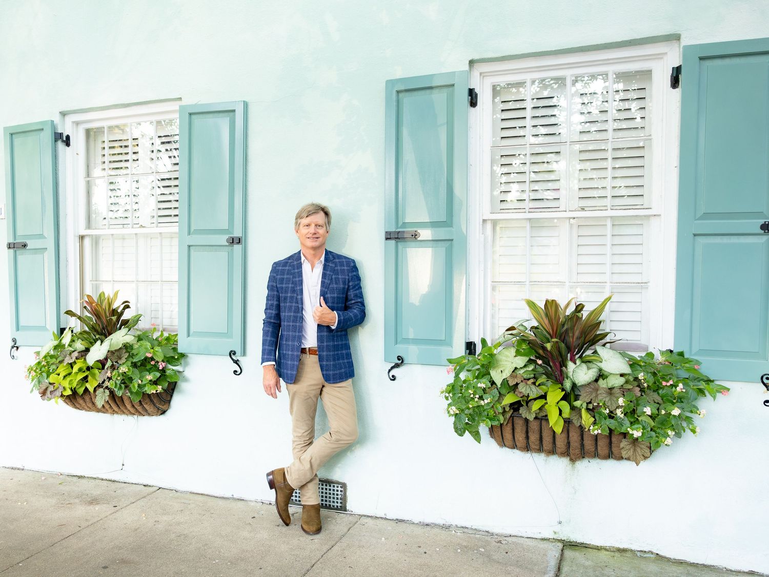 Man in blue blazer leaning against a light blue building with teal shutters and lush window boxes.