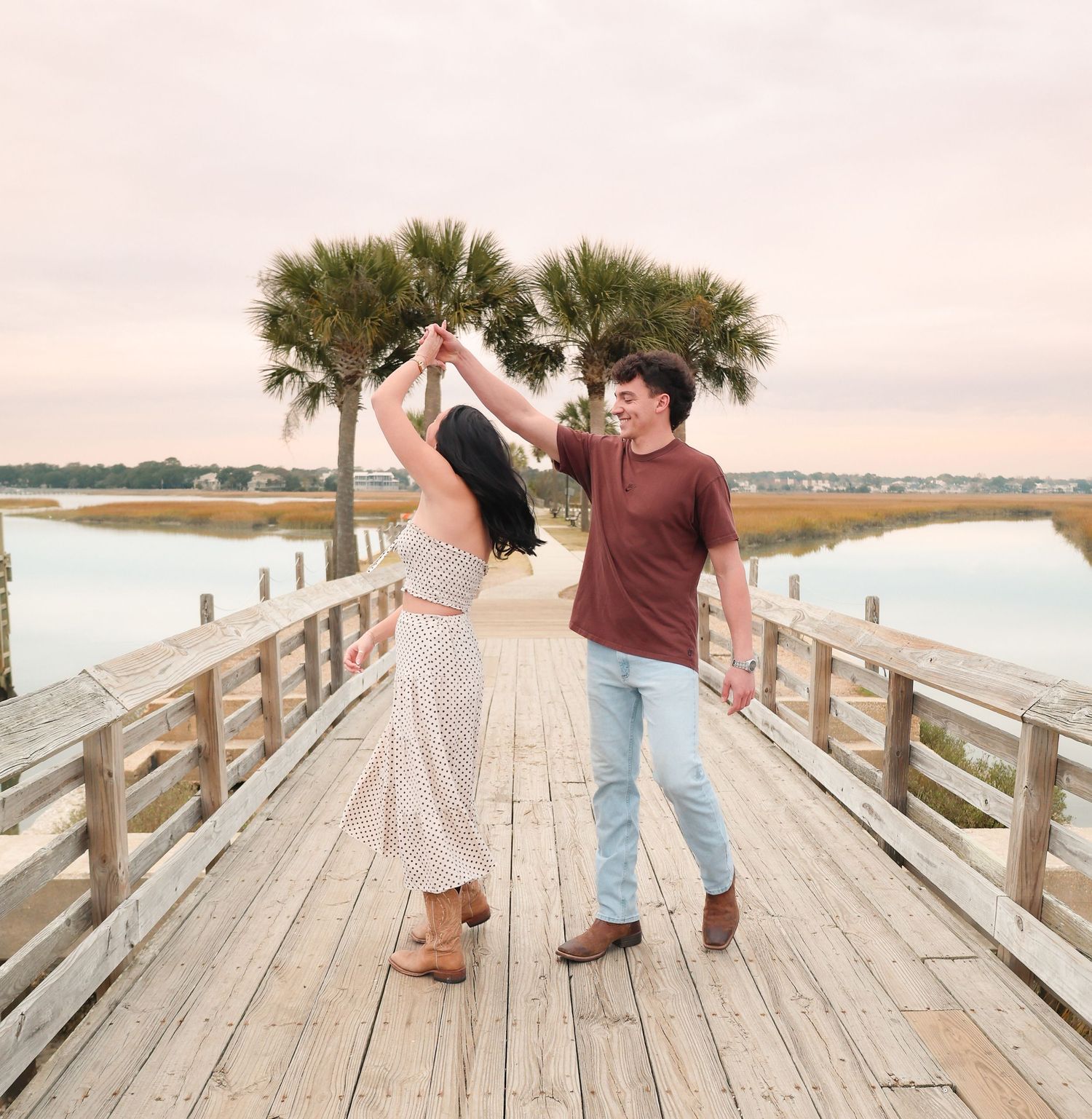 A couple dances on a wooden pier surrounded by palm trees and a serene waterway at sunset.