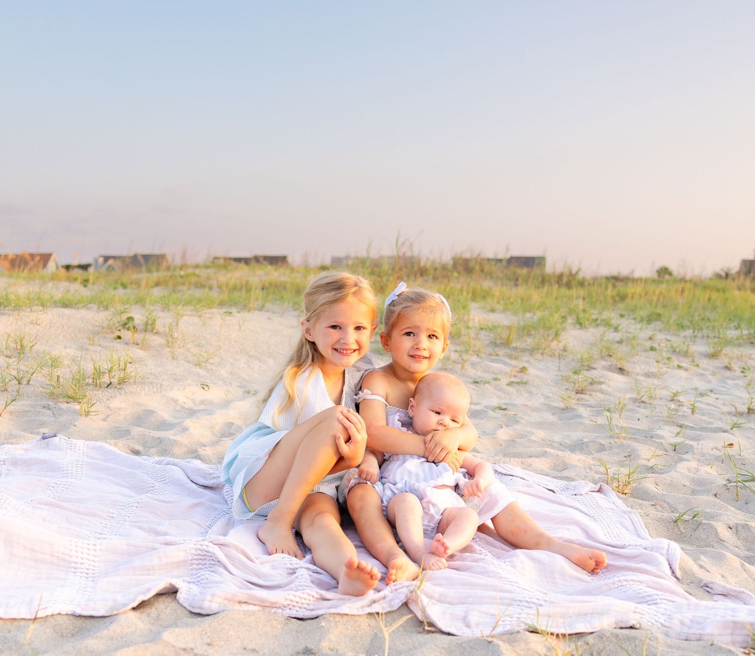 Two young girls and a baby sibling sit together on a blanket at a sandy beach during golden hour.