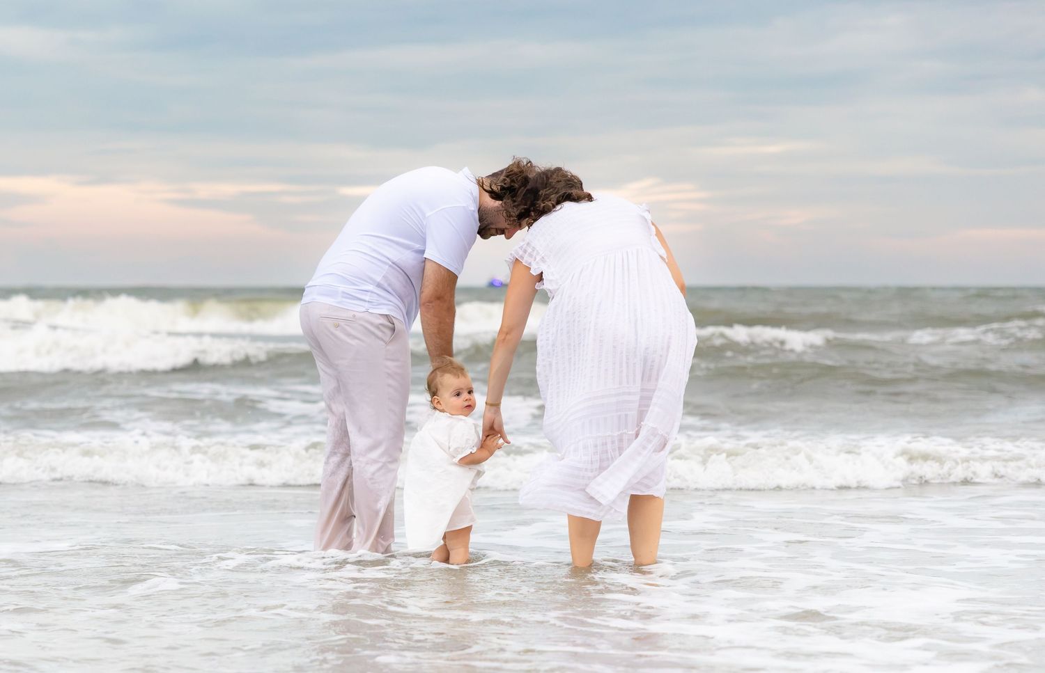 Two adults help a baby walk along a sandy beach with ocean waves in the background on a cloudy day.