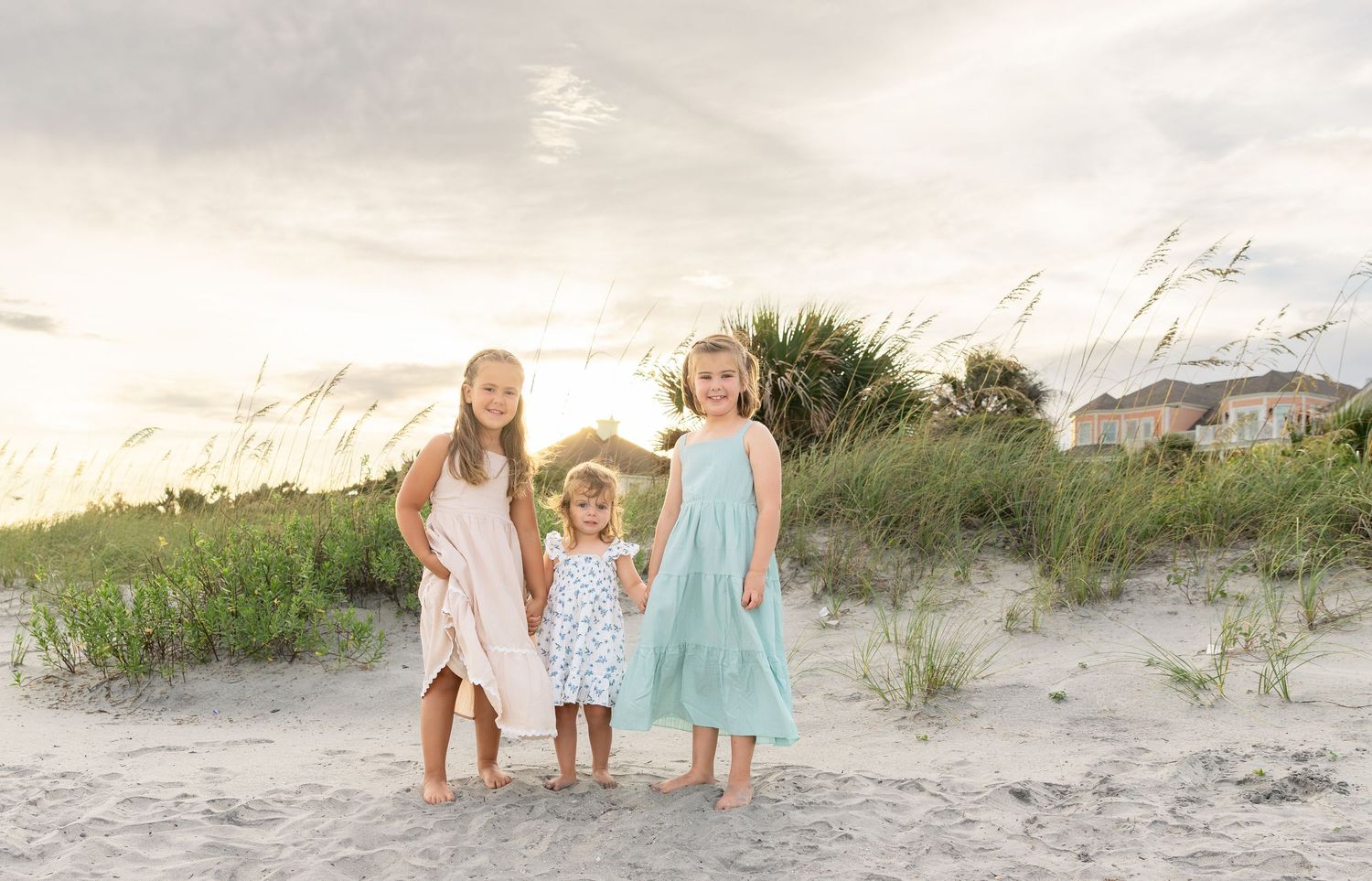 Three young girls in pastel dresses pose on a sandy beach at sunset with sea oats and coastal homes in the background.