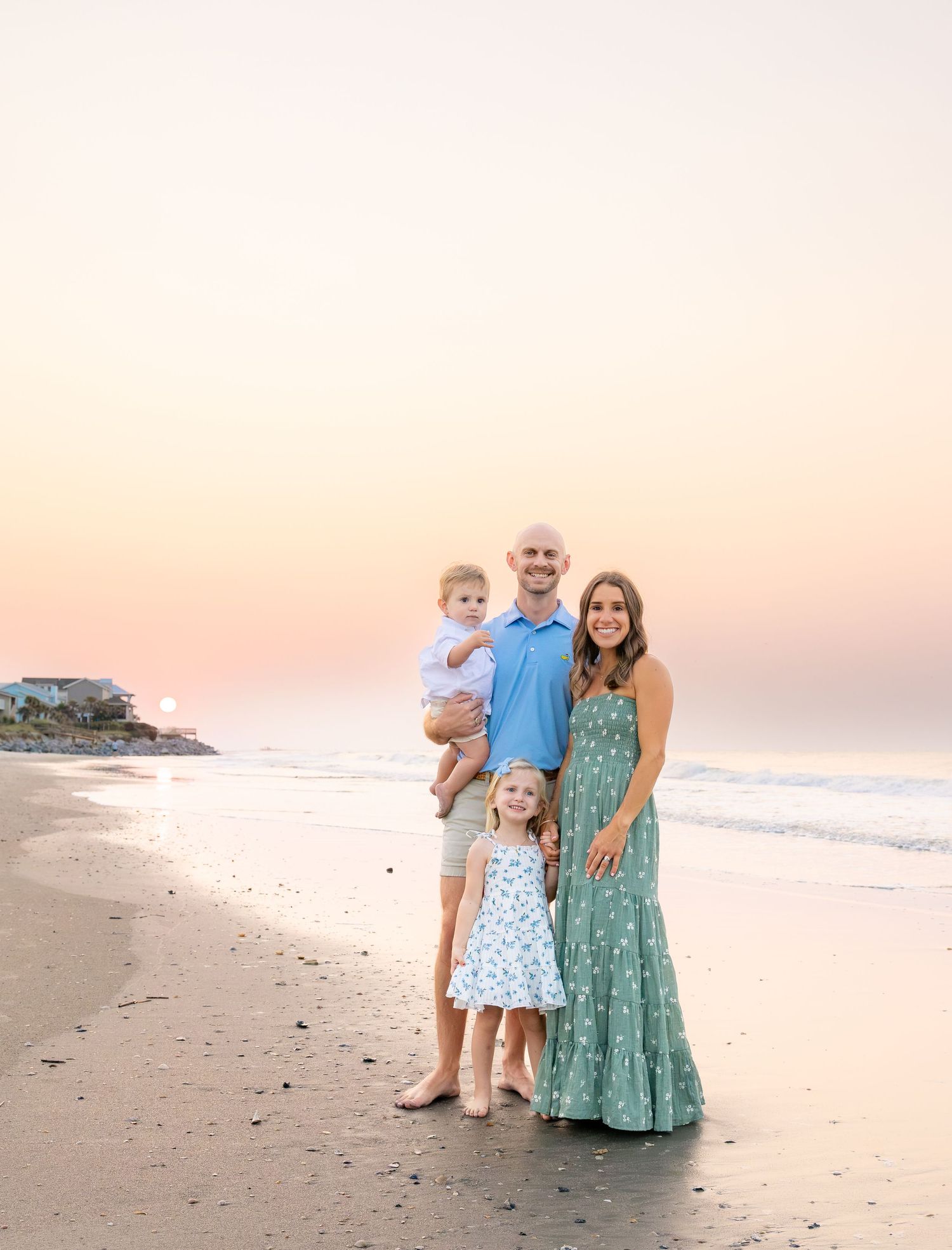 Family of four posing on a beach at sunset, wearing coordinated blue and green outfits, with calm ocean waves in the background.