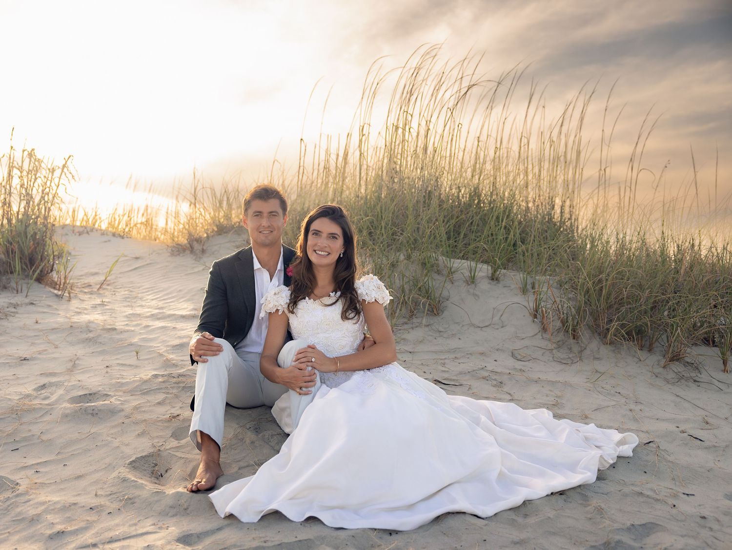 Bride and groom sitting on sandy beach dunes at sunset, woman in white wedding dress, man in gray suit.