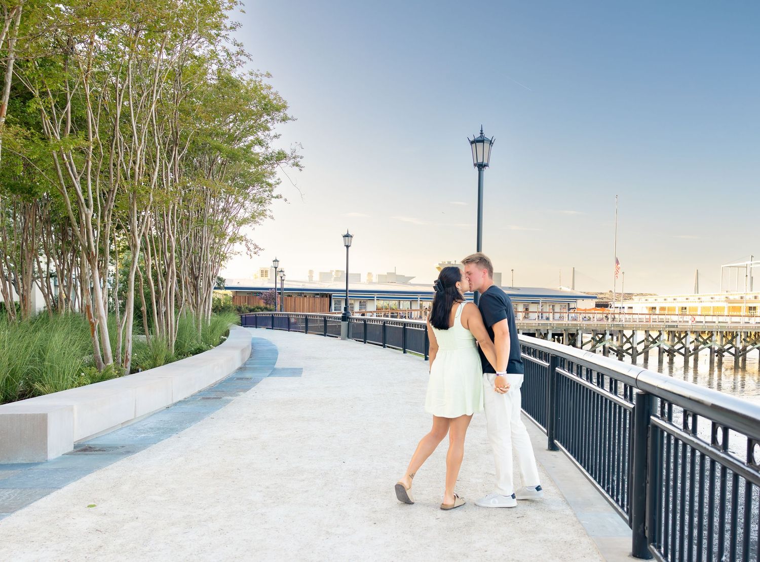 Couple sharing a romantic kiss on a waterfront promenade with trees and a pier in the background.