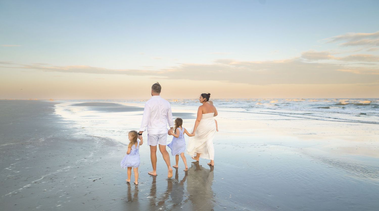 A family of four walks hand-in-hand along a serene beach at sunset in white and blue outfits.