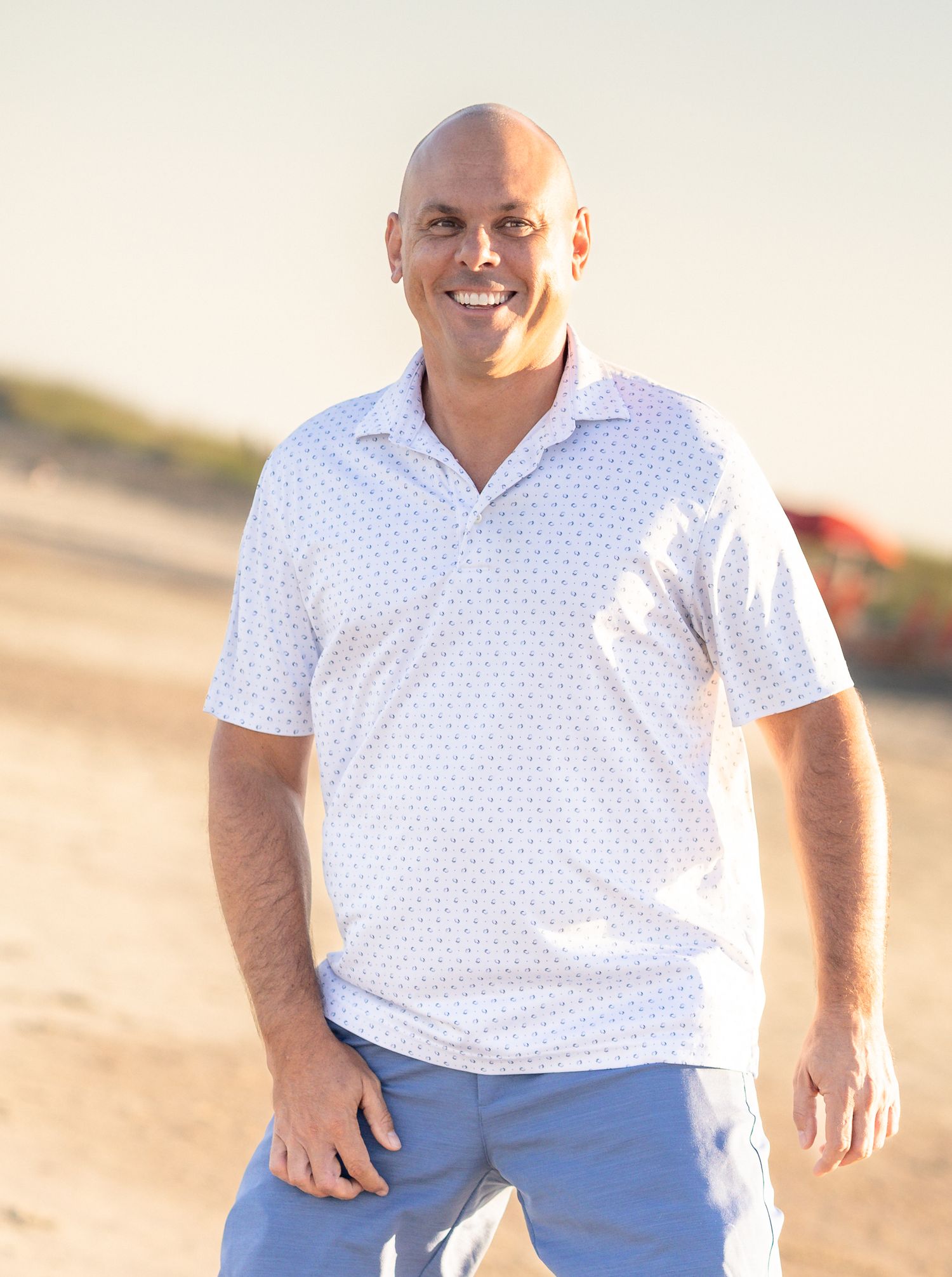 Smiling bald man in white patterned polo shirt and blue pants standing on a sunny beach.