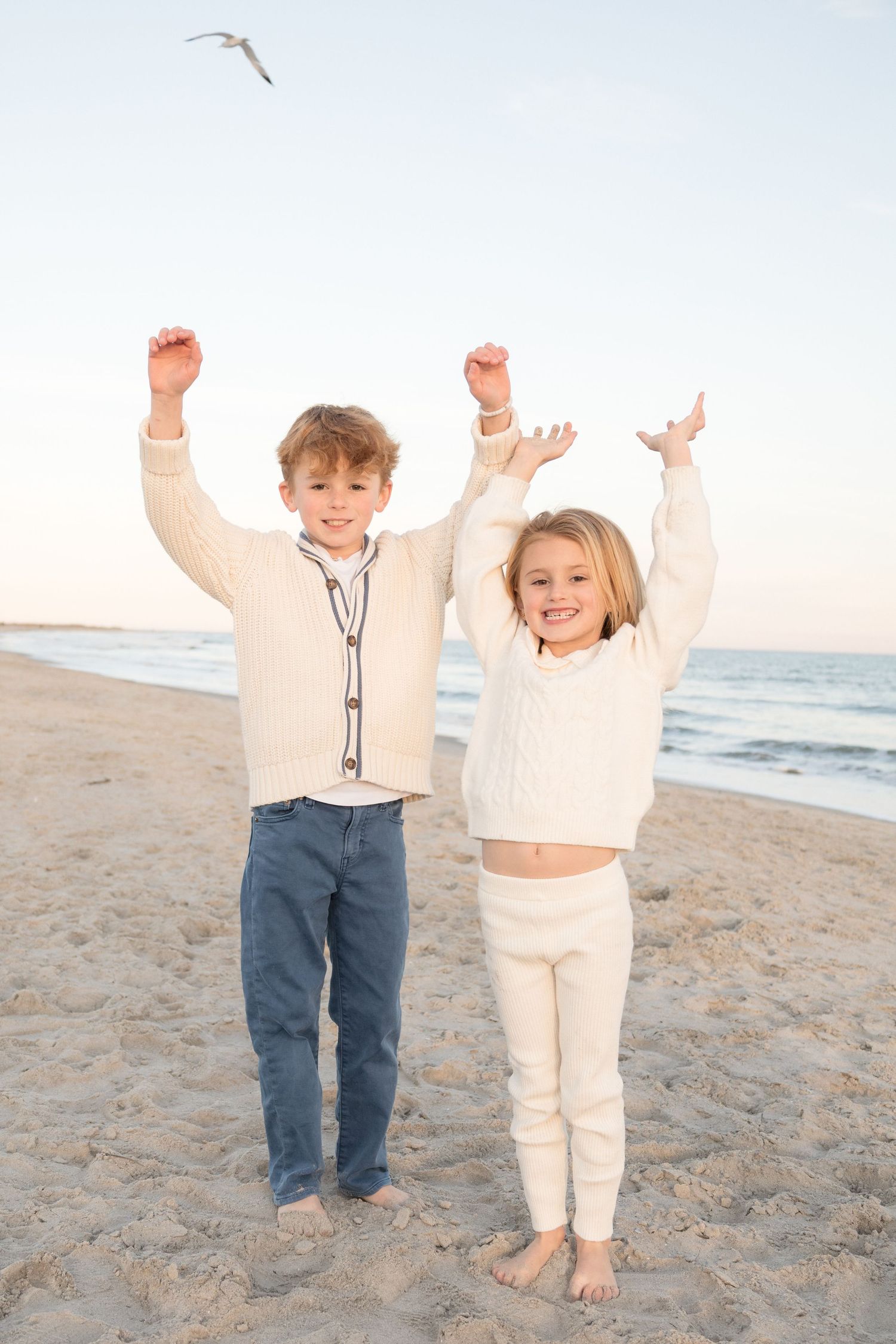 Two children jumping with joy on a sandy beach at sunset with ocean waves in the background.