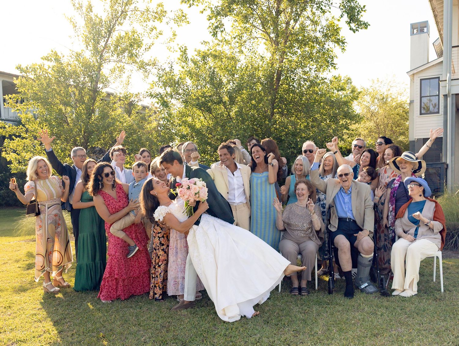 Wedding party guests cheer as bride and groom share romantic kiss on sunny outdoor lawn.