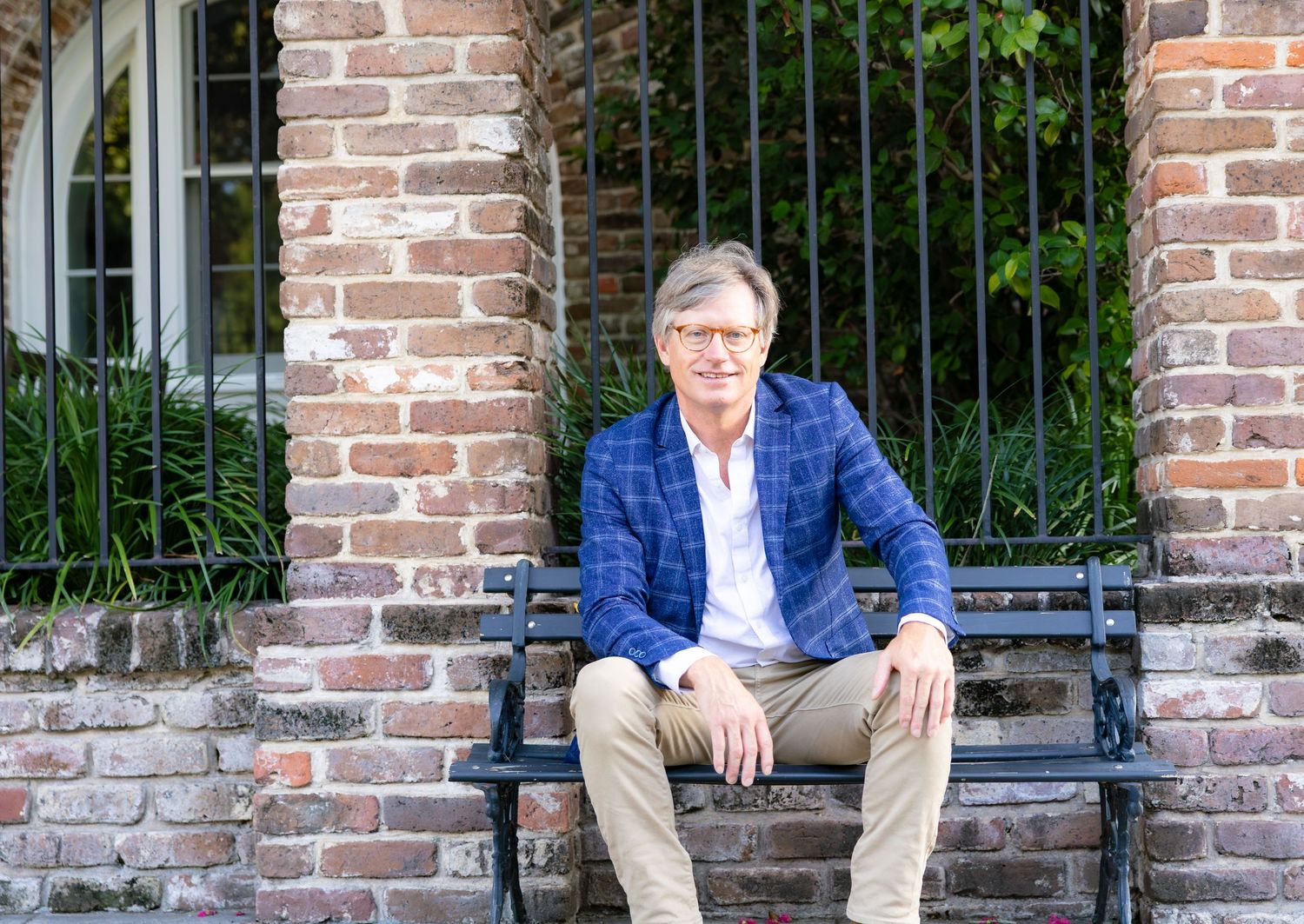 Man in blue plaid blazer sitting on black bench against rustic brick wall with iron fence outdoors.