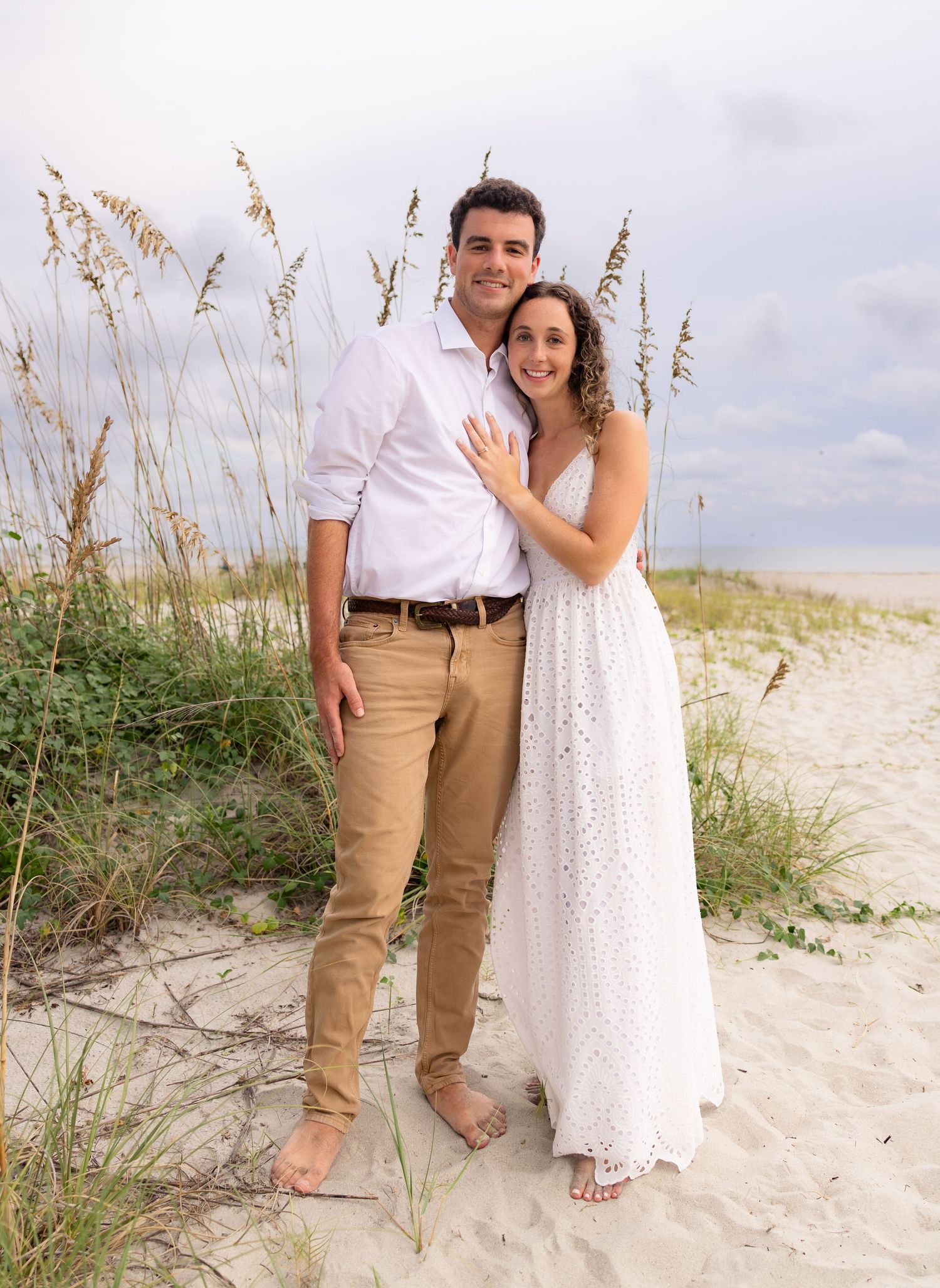 Happy couple posing together on a sandy beach with sea oats, woman in white dress and man in khaki pants.