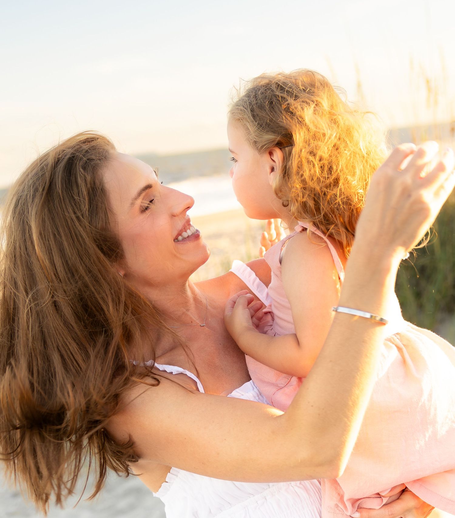 A smiling woman lifts and embraces a young girl with curly hair on a sunny beach, both wearing light summer clothing.