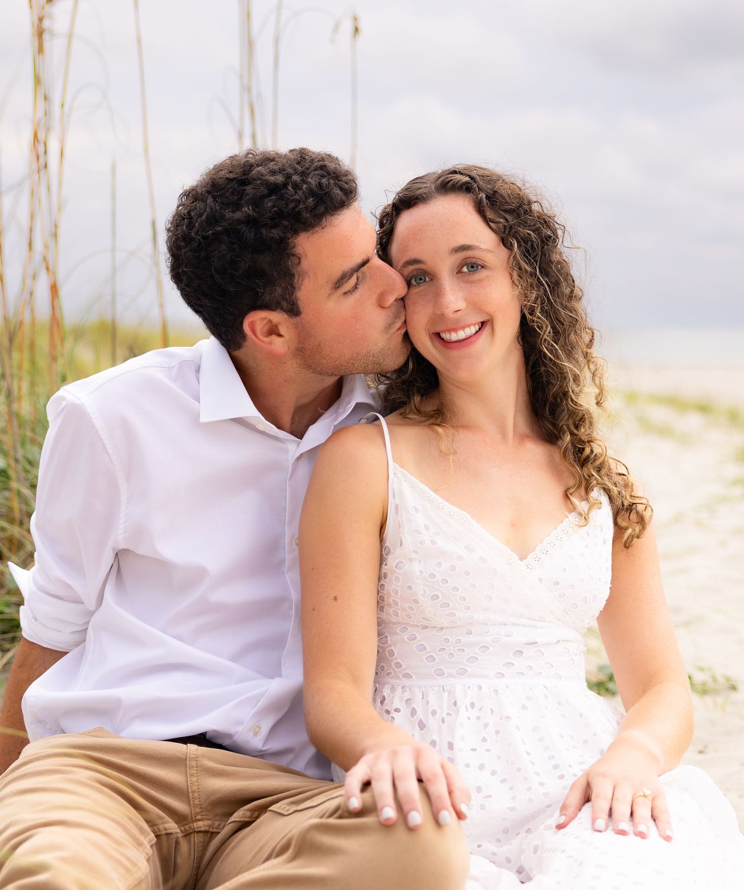 Couple sharing a romantic moment on a sandy beach, man kissing woman's cheek as she smiles in white dress.