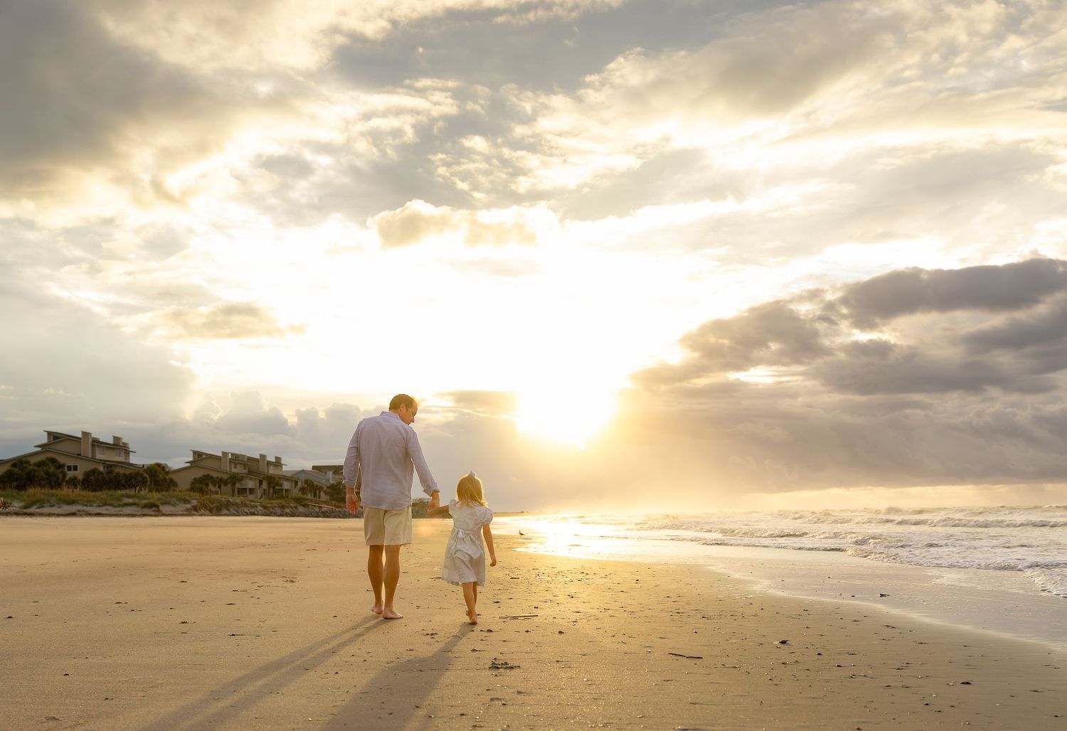 Adult and child walking hand-in-hand on a sandy beach at golden sunset with dramatic clouds overhead.