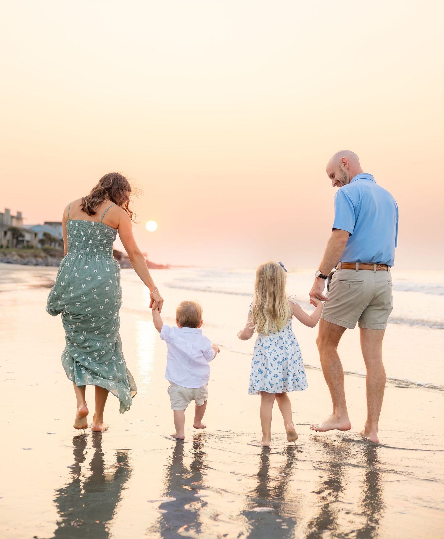 Family of four walking hand-in-hand on a beach at sunset, two parents with toddler and young child.