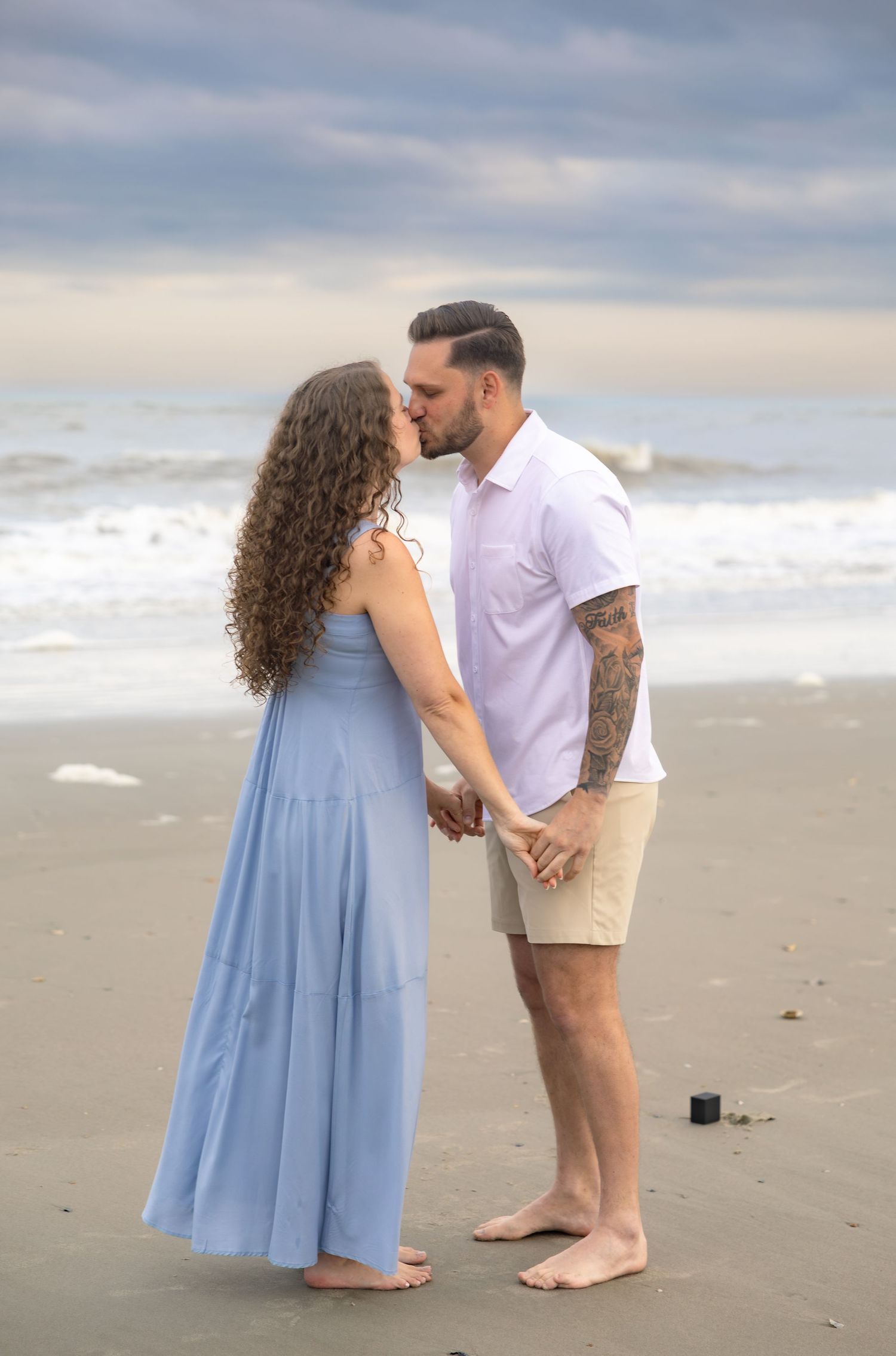 Couple shares romantic kiss on sandy beach while holding hands with ocean waves behind them.