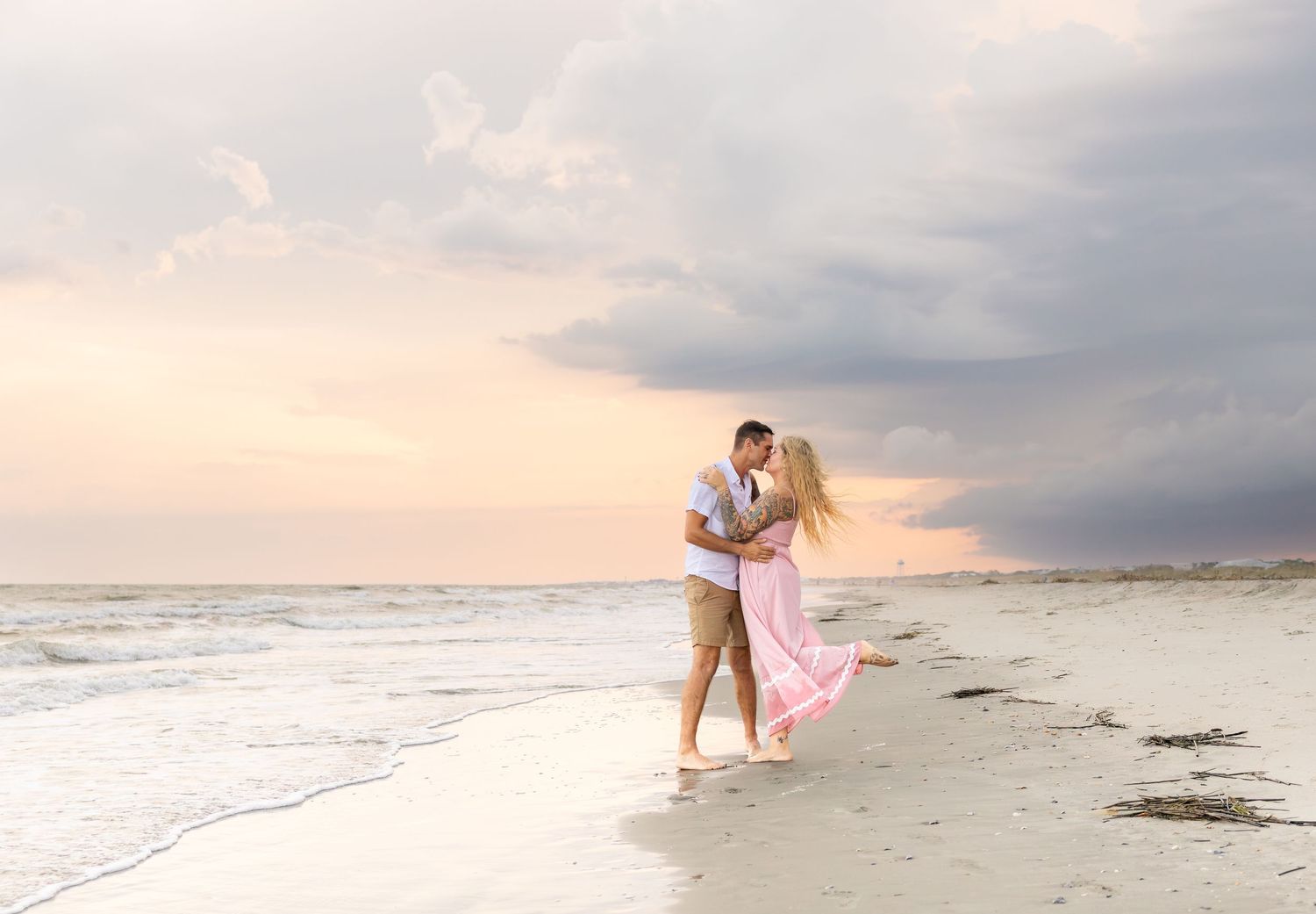 Couple sharing a romantic kiss on a sandy beach at sunset, woman in flowing pink dress.