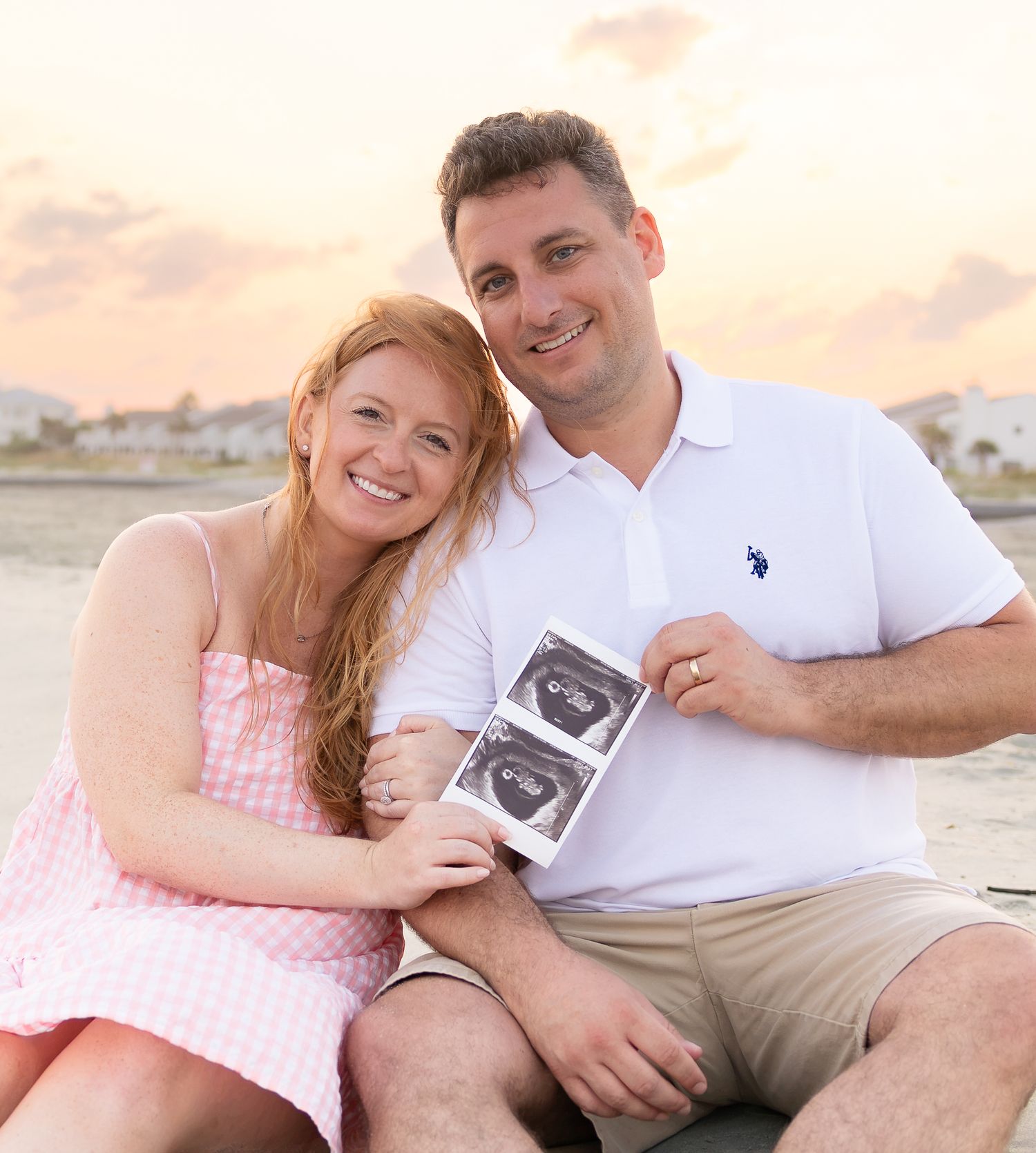 Happy couple sitting on beach holding ultrasound photos, announcing pregnancy at sunset.