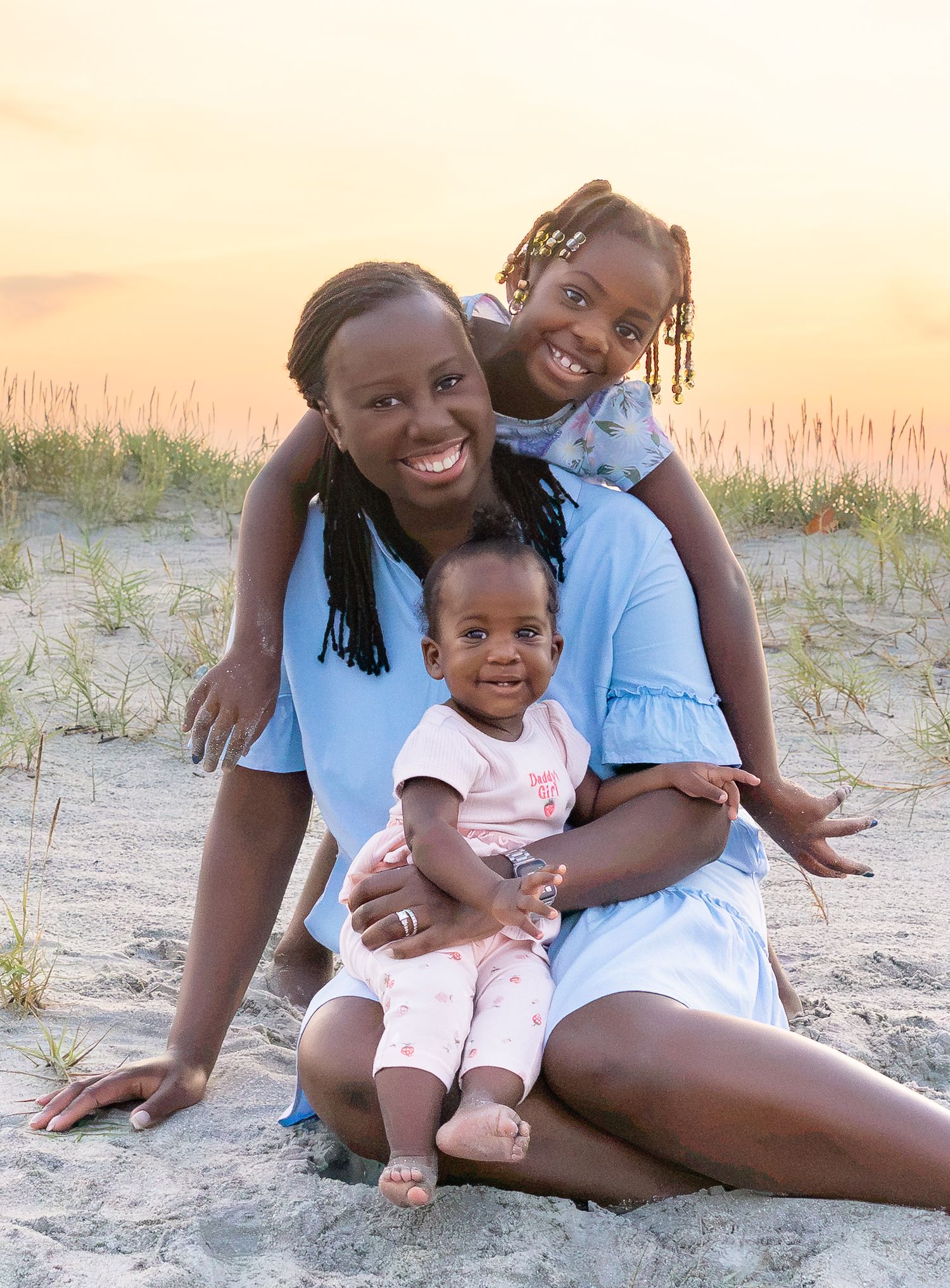 A mother and two daughters smile together on a sandy beach at sunset, wearing matching light blue outfits.