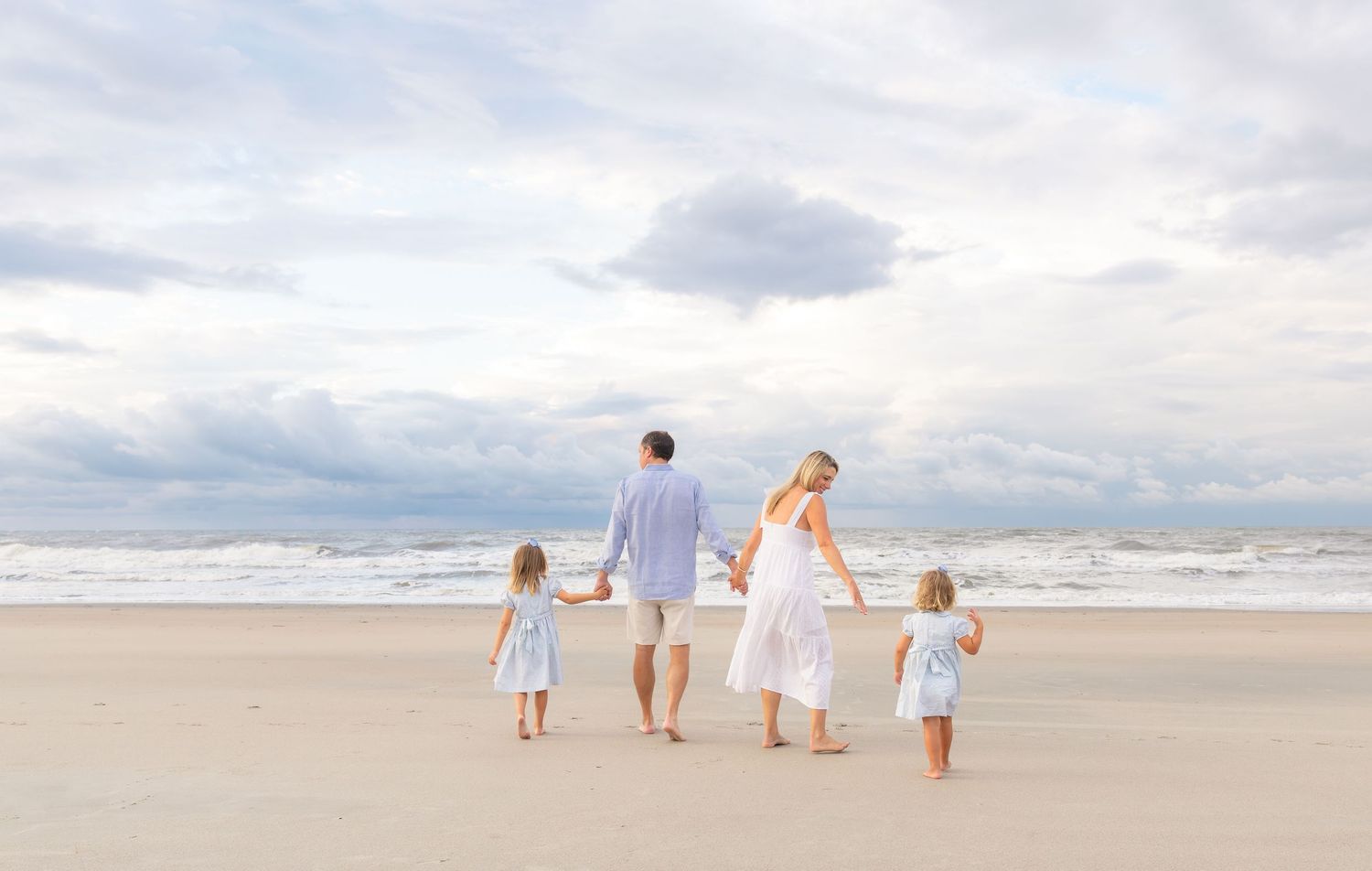 Family of four walking hand-in-hand on a sandy beach under a cloudy sky.