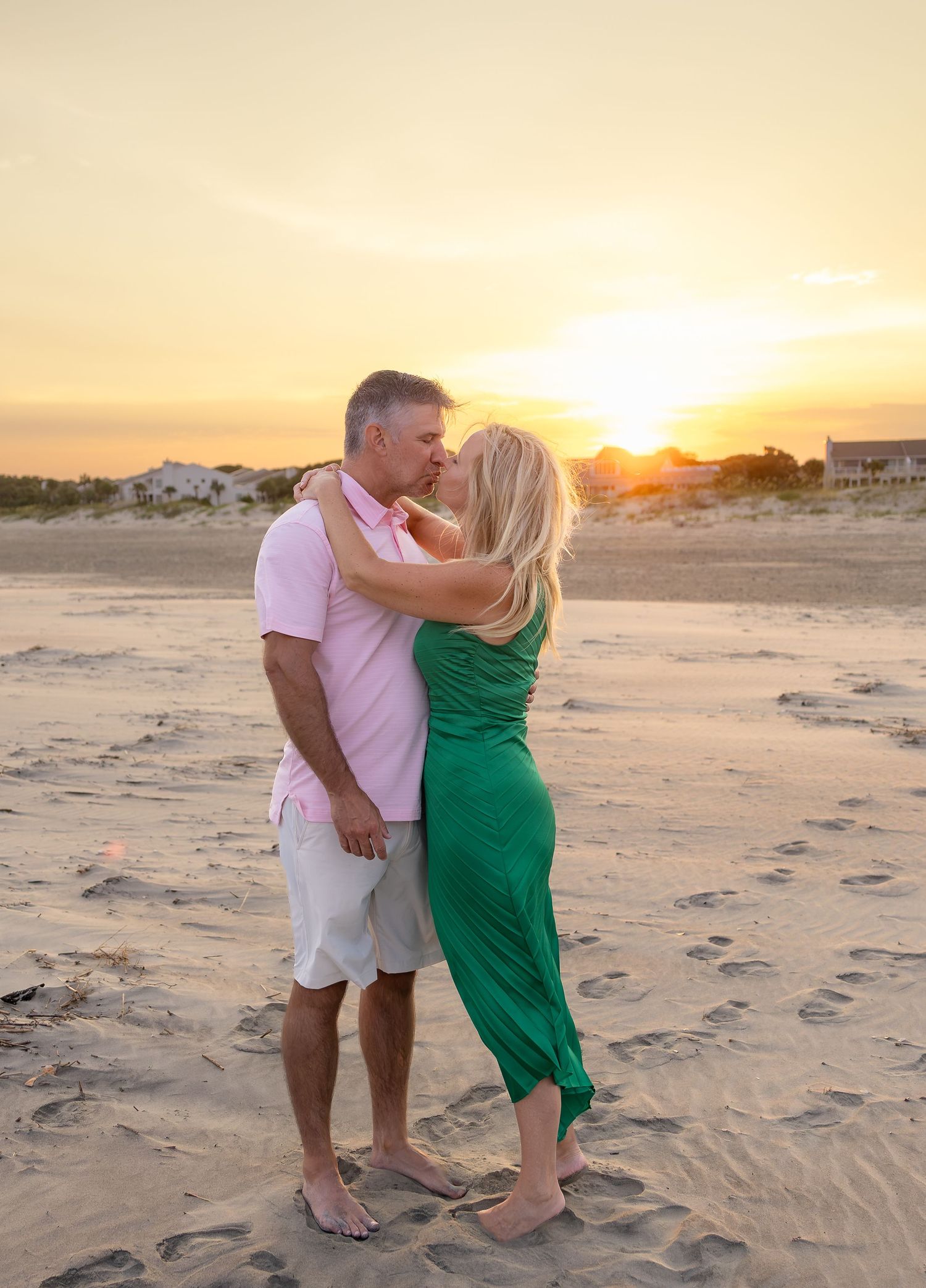 Couple in pink shirt and green dress embracing barefoot on a sandy beach at sunset with golden sky and coastal houses behind.
