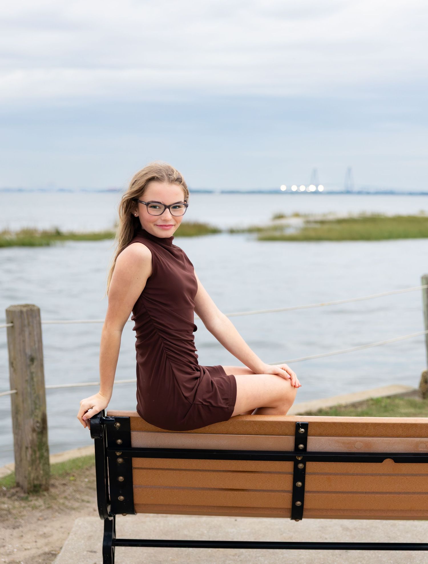 Young woman in glasses and a burgundy dress sits on a wooden waterfront bench overlooking calm water and marshland under a cloudy sky.