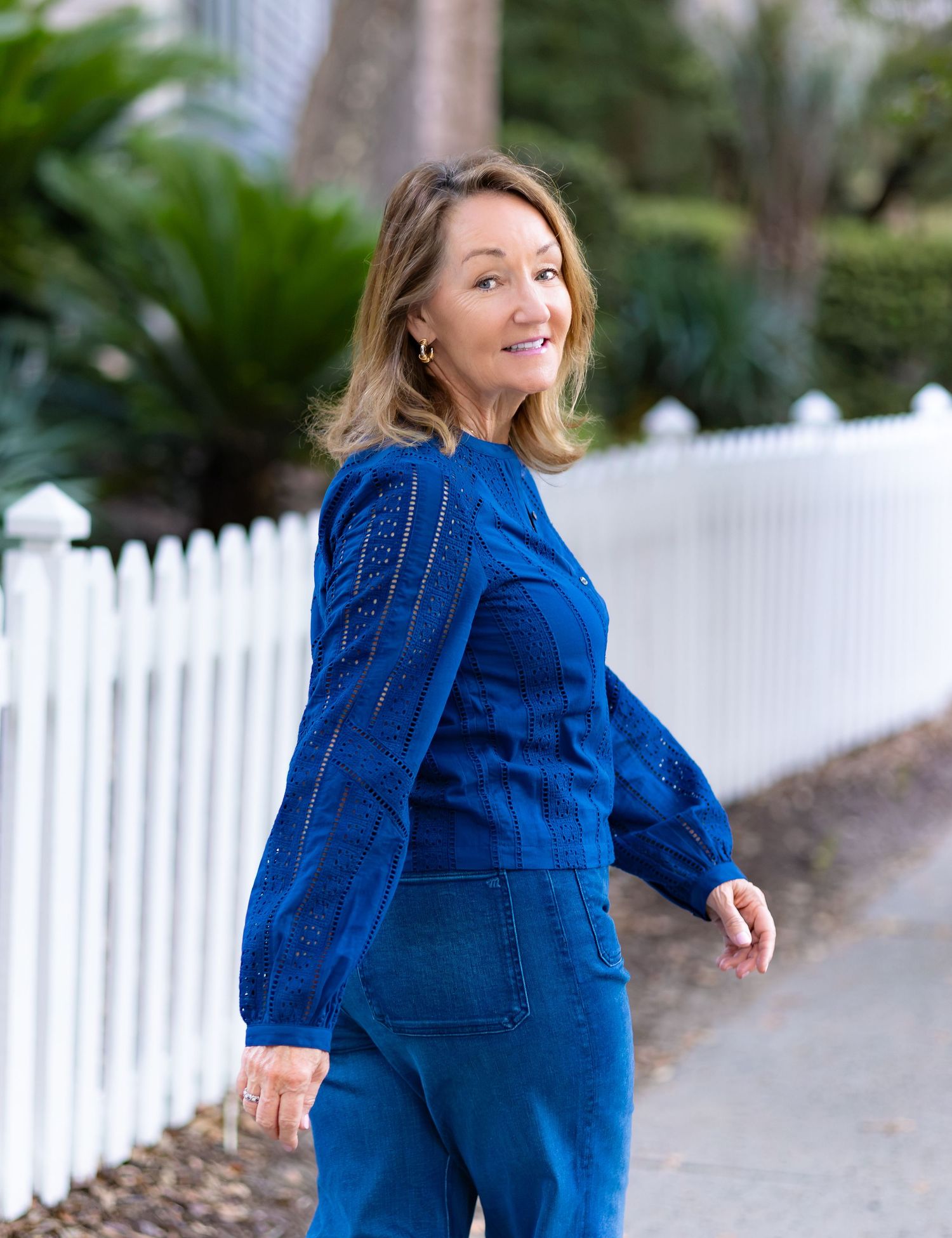 Woman in cobalt blue lace top and denim pants poses on a tree-lined sidewalk near a white picket fence.