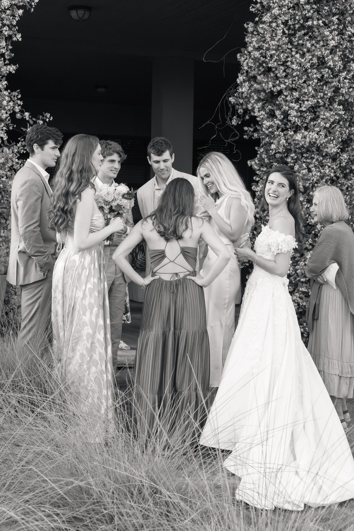 Black and white photo of a smiling bride in a flowing off-the-shoulder gown chatting with wedding guests outdoors near a flowering hedge.