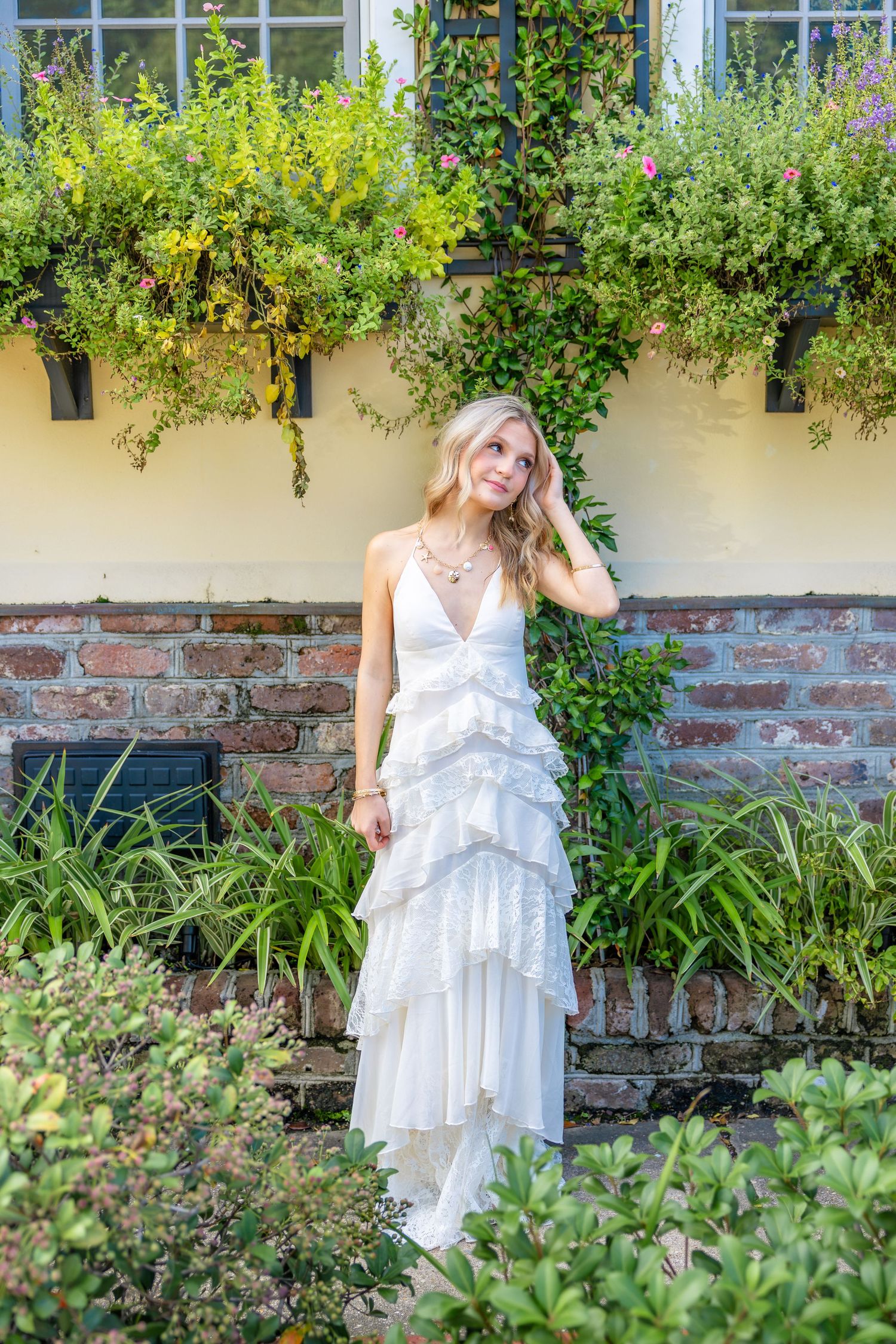 Woman in a white tiered ruffle dress poses with her hand in her hair before a brick wall with lush green plants and flowering window boxes.