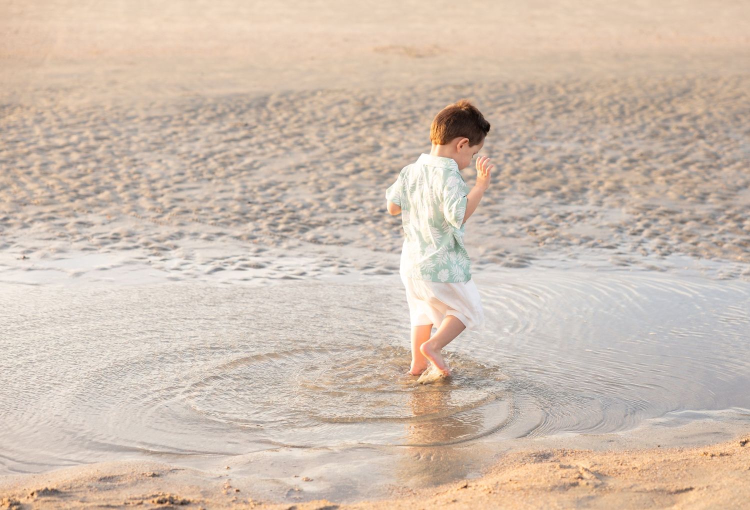 Young child wading in shallow ocean waves on a sandy beach, wearing a light blue outfit.