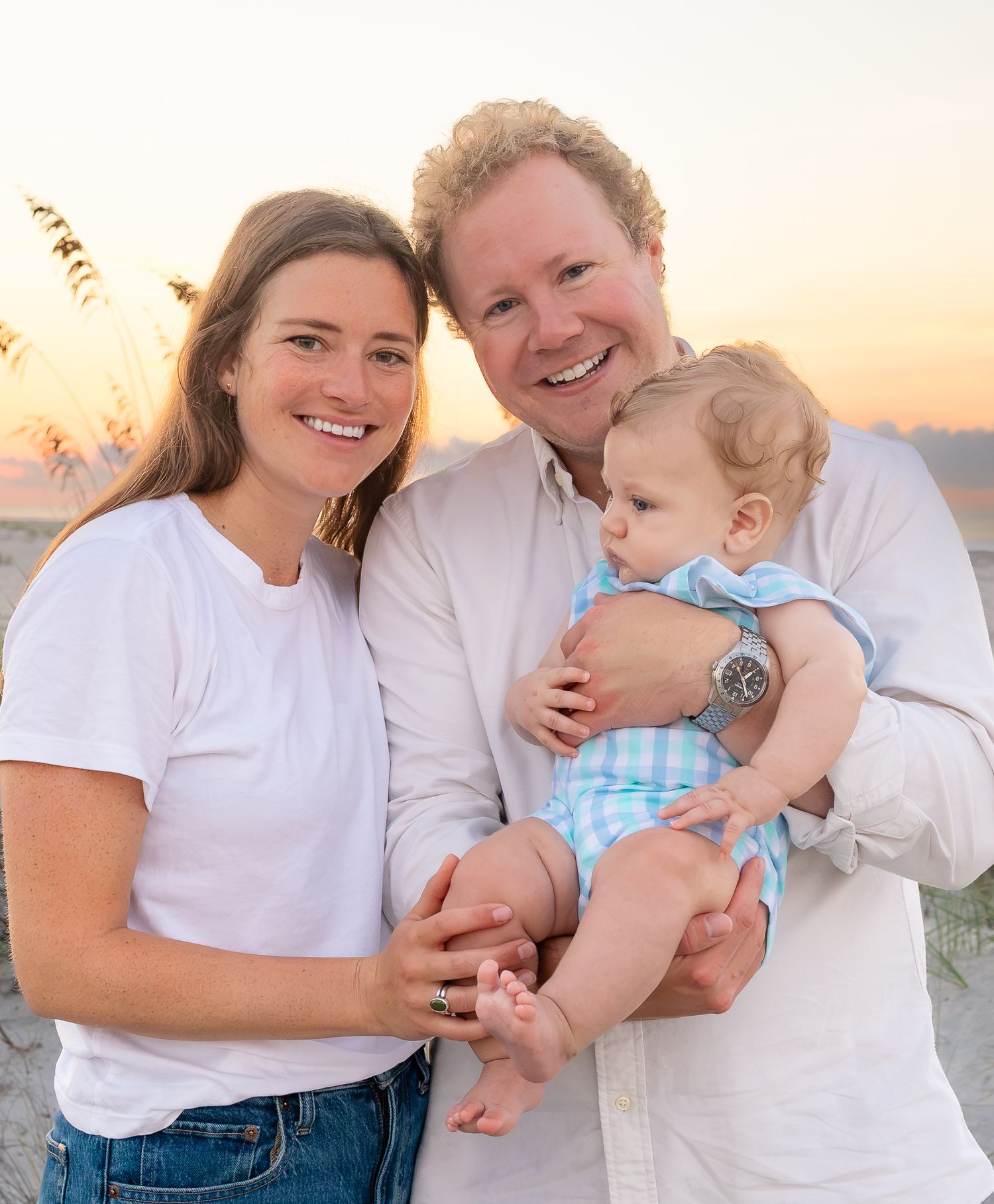 A smiling young family with a baby poses on a sandy beach at golden hour sunset.