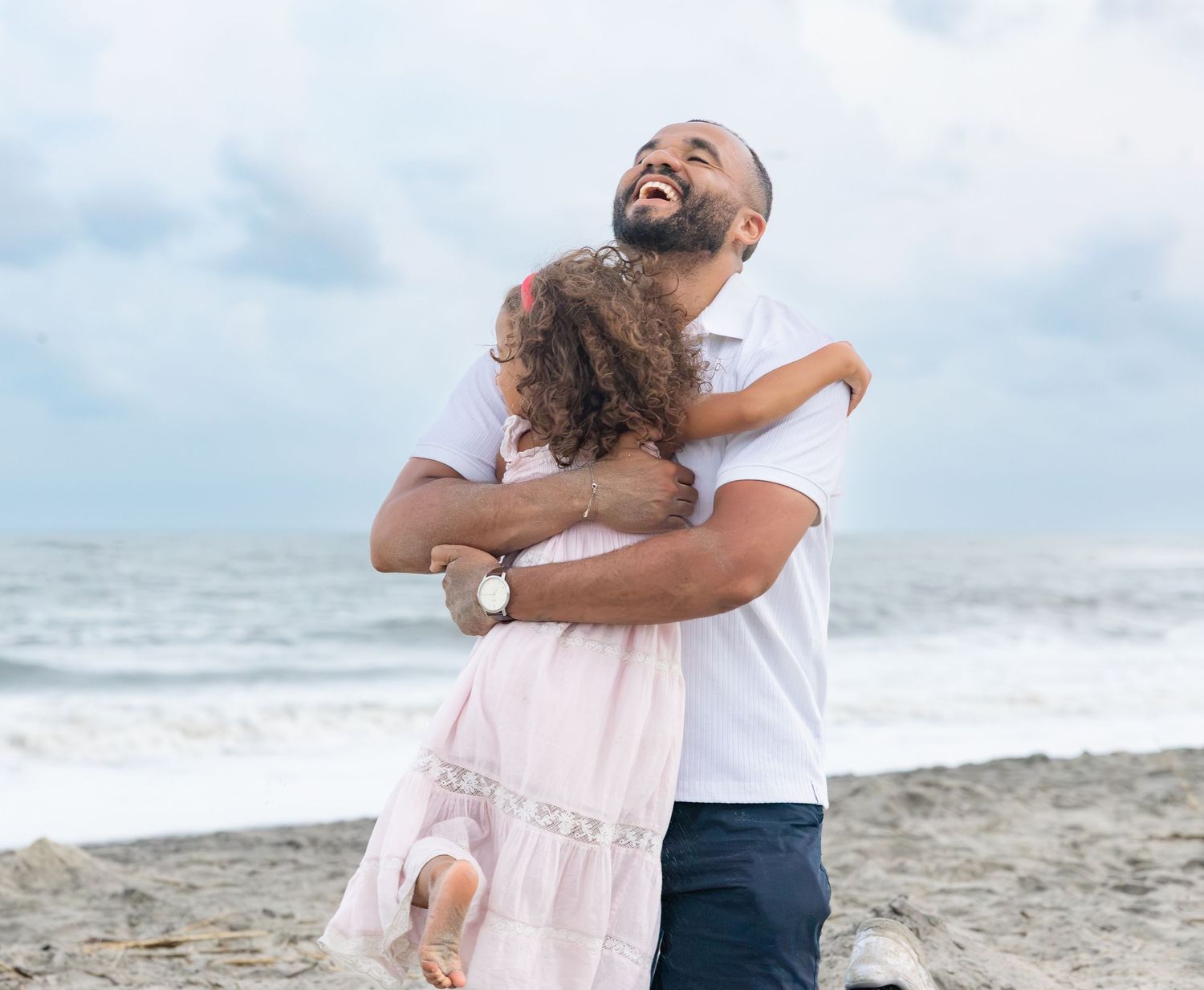 A joyful man hugging a young girl on a beach, both smiling, with ocean waves in the background.