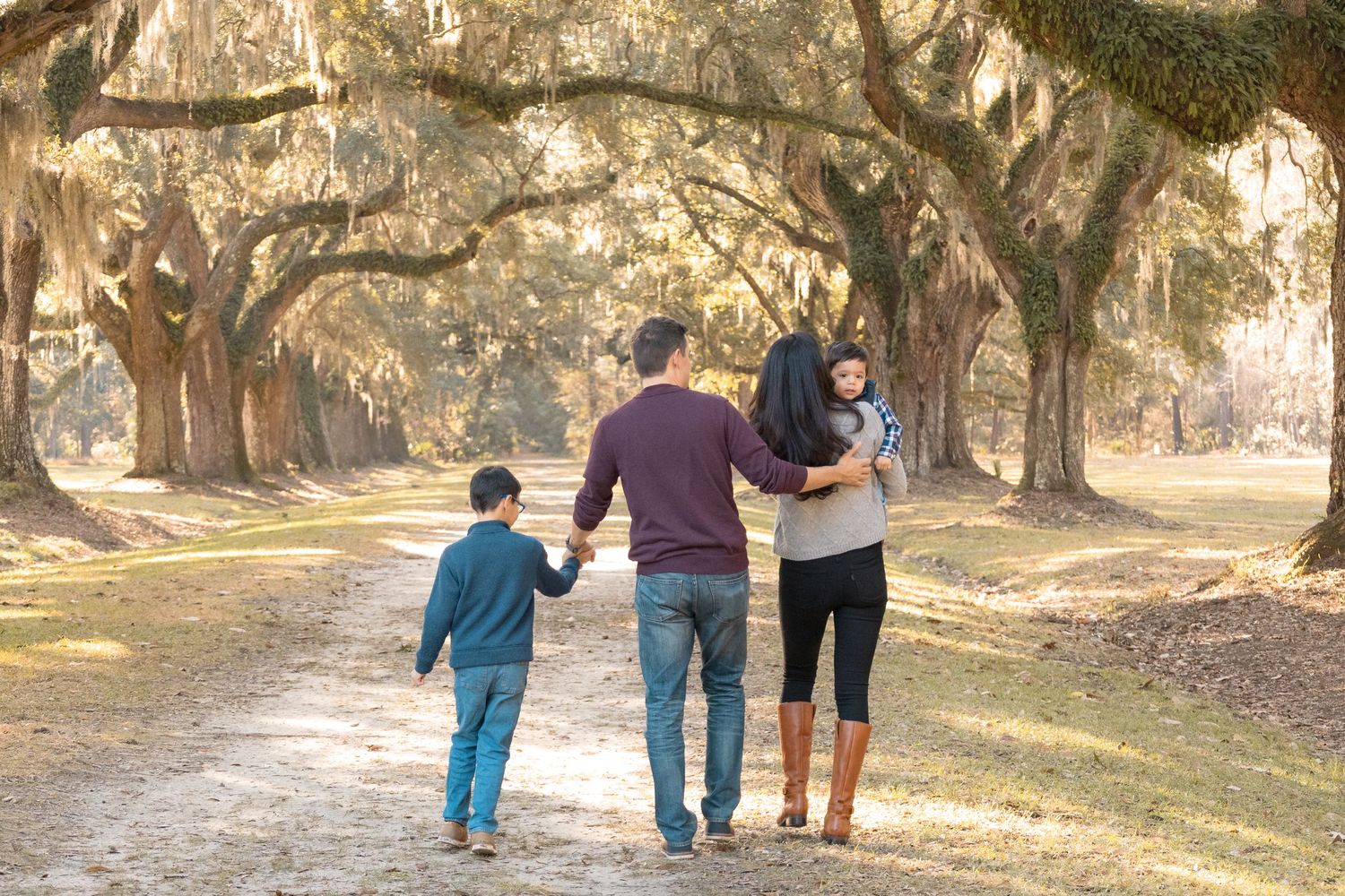 Family of four walks hand in hand down a sunlit dirt path beneath live oak trees draped in Spanish moss.