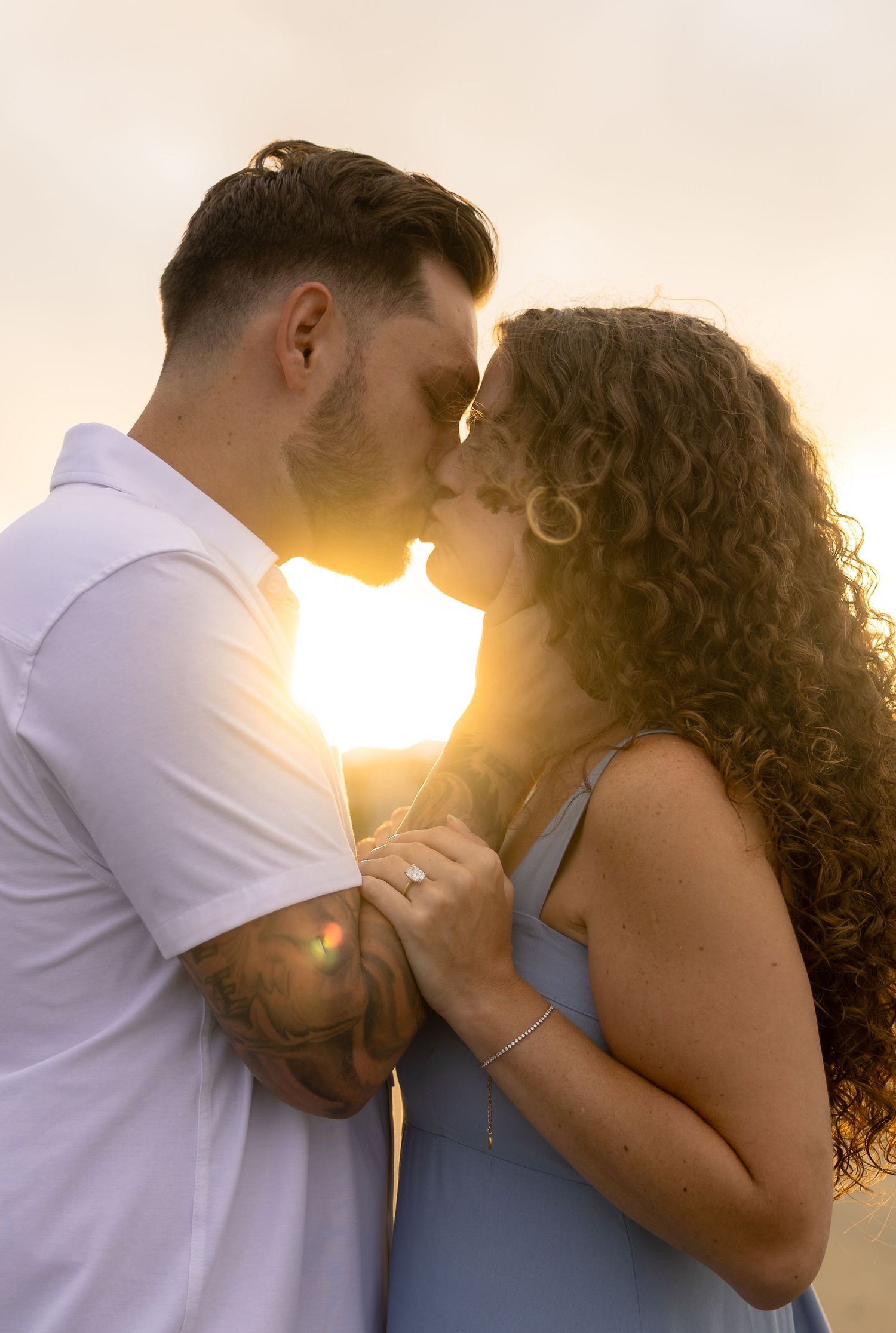Romantic couple about to kiss at sunset on the beach, woman wearing engagement ring, backlit by golden light.