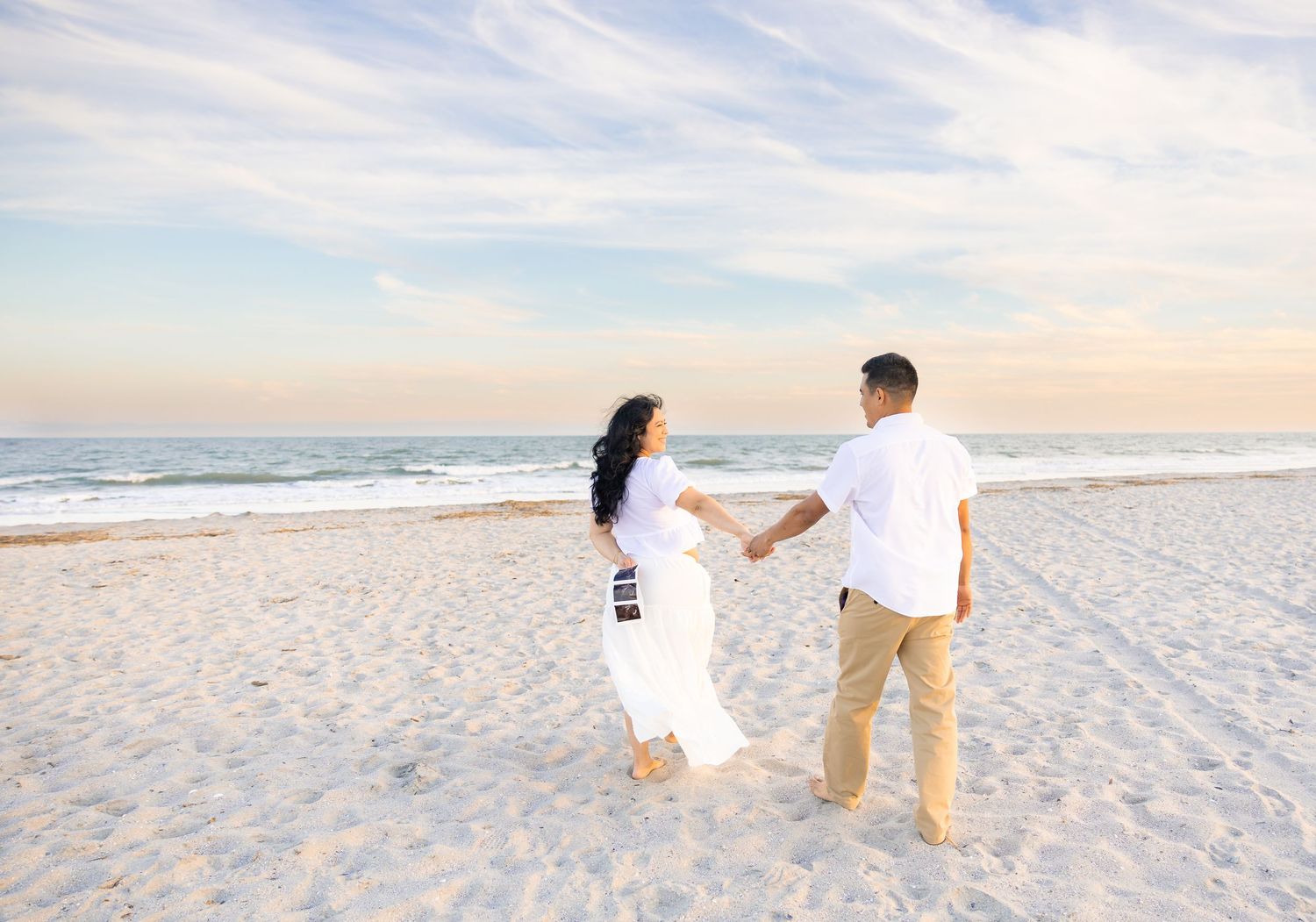 Couple holding hands on a white sandy beach at sunset, woman in white dress, man in white shirt and khaki pants.