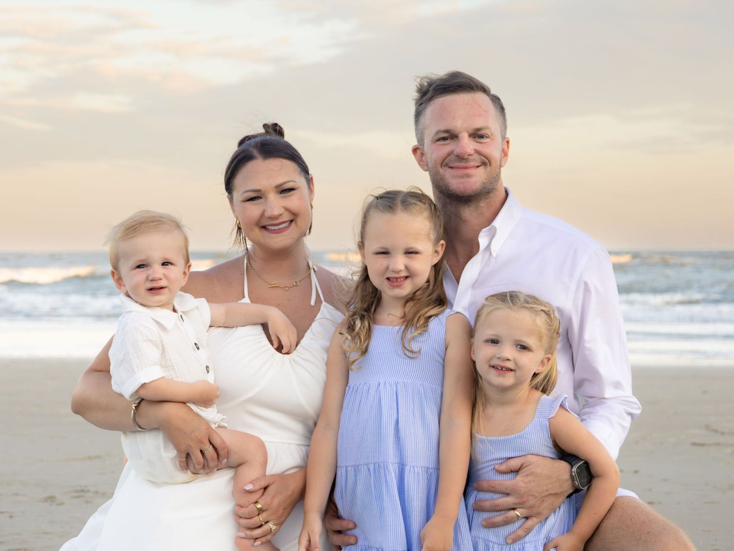 Family of five smiling together on a sandy beach at sunset, parents holding young children in white and blue outfits.