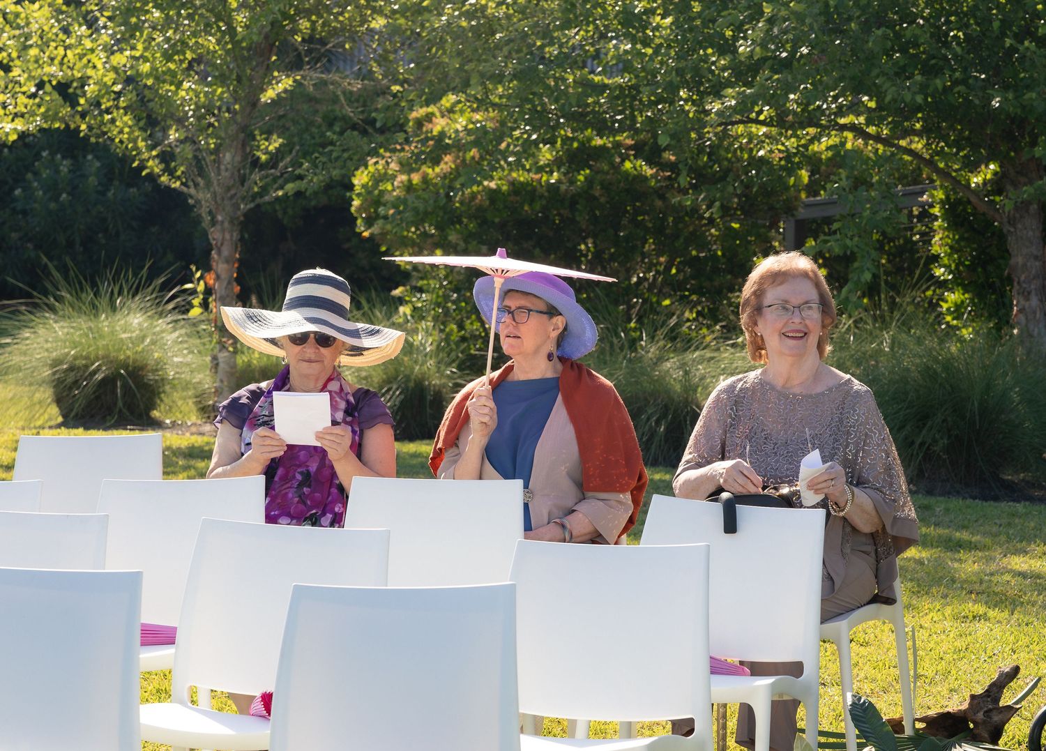 Three women wearing elegant hats sit in white chairs at an outdoor garden ceremony.