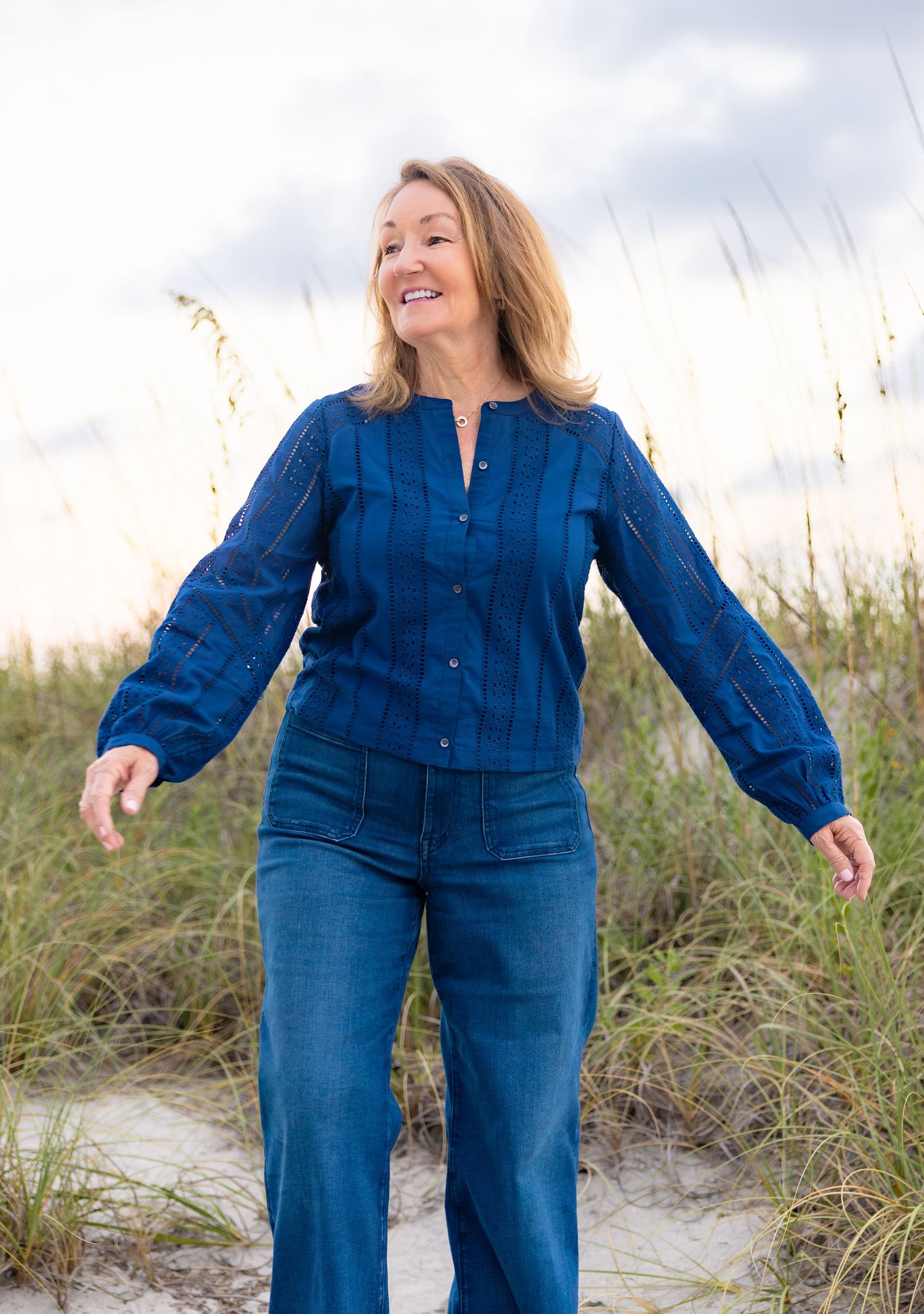 Smiling woman in blue blouse and jeans walking on sandy beach dunes with sea oats in background.