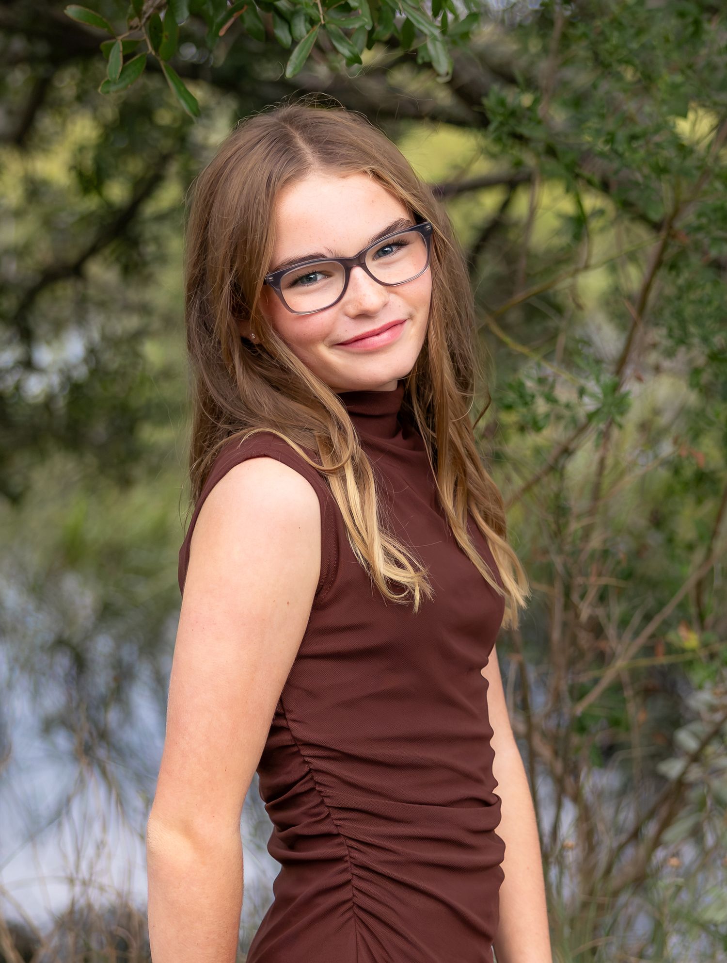 Young girl with glasses and brown dress smiling outdoors near trees and water.