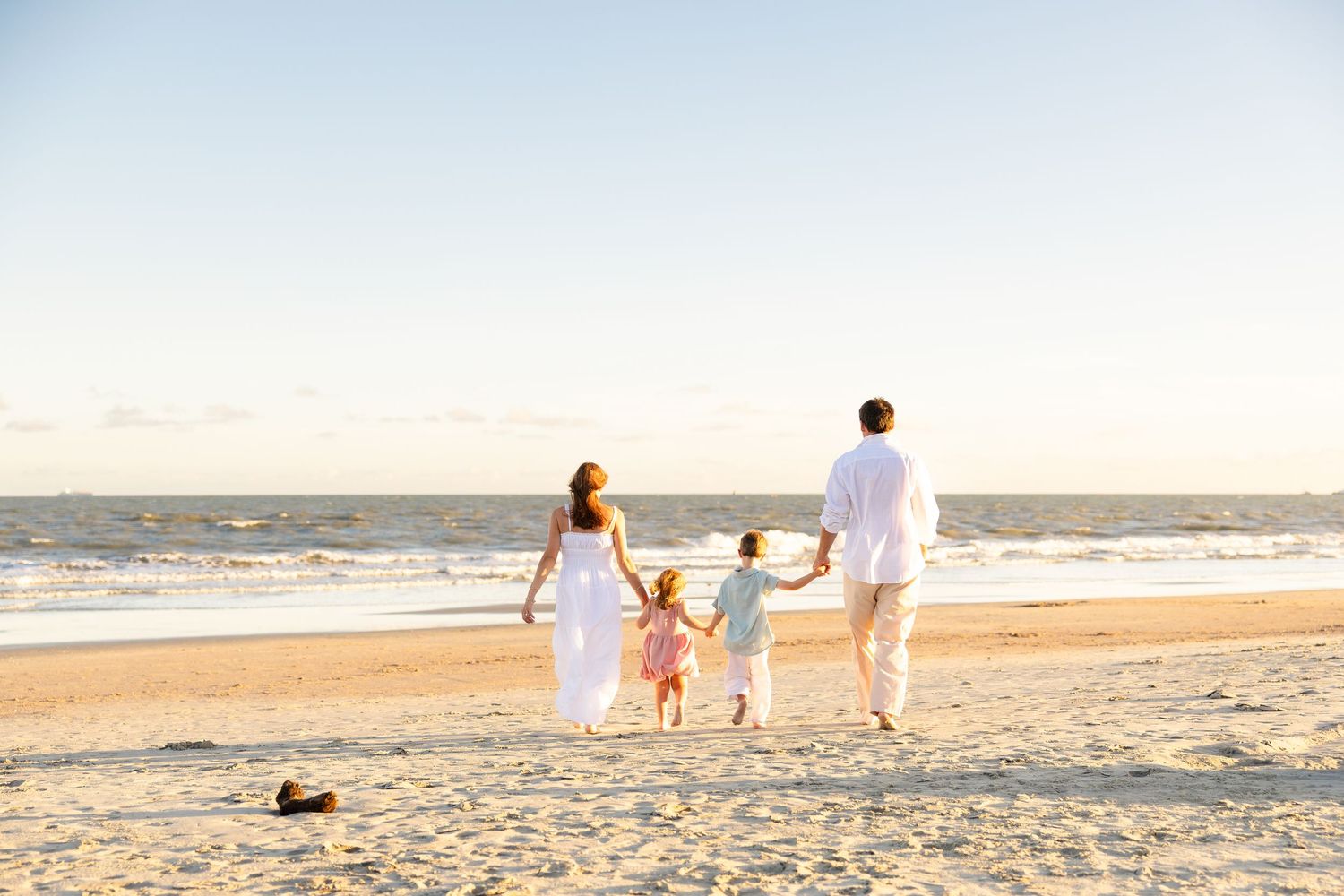 Family of four holding hands and walking along a sandy beach toward the ocean at sunset, with a woman in a white dress.
